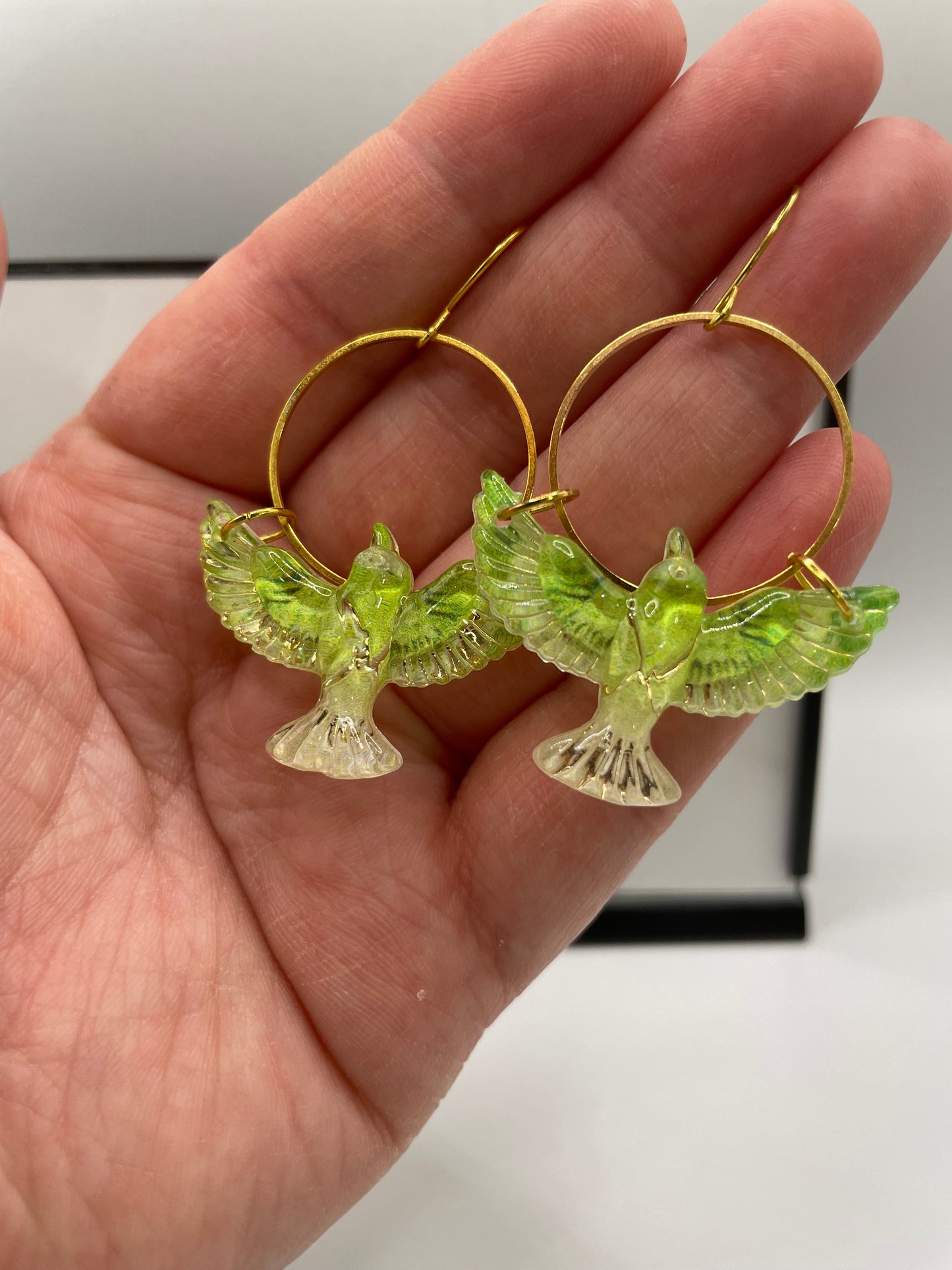 Hand holding gold hoop earrings with green bird-shaped charms against a neutral background
