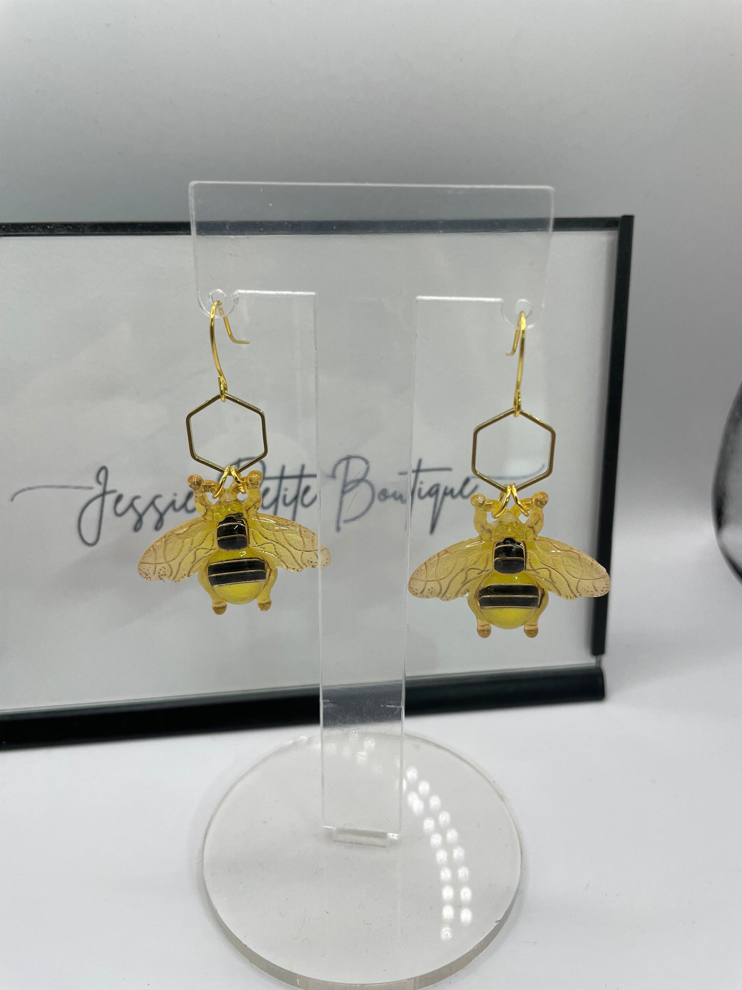 Gold bee-shaped earrings on a clear stand with a branded card in the background.