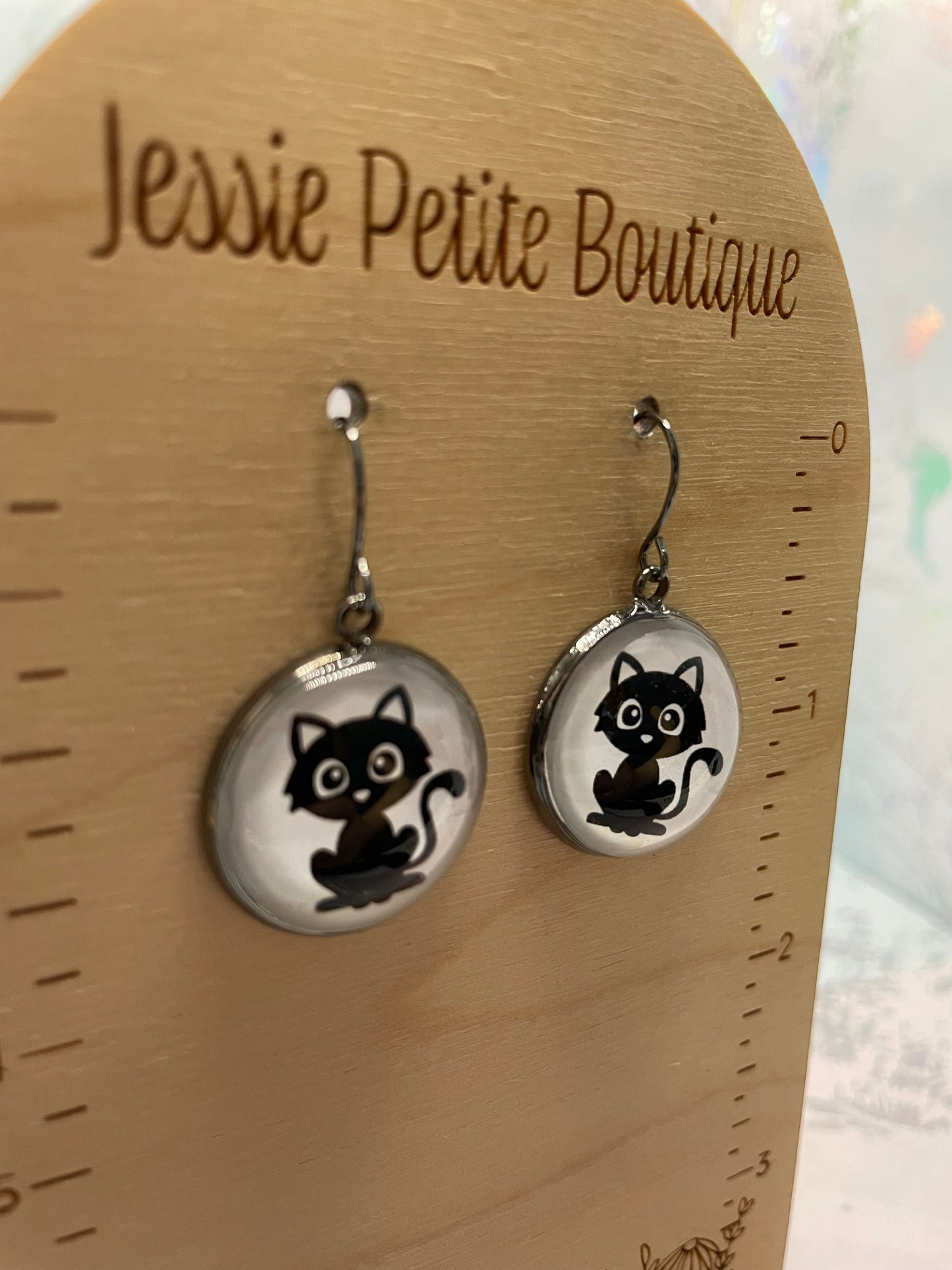 Cat-themed earrings on a wooden ruler with 'Jessie Petite Boutique' branding.