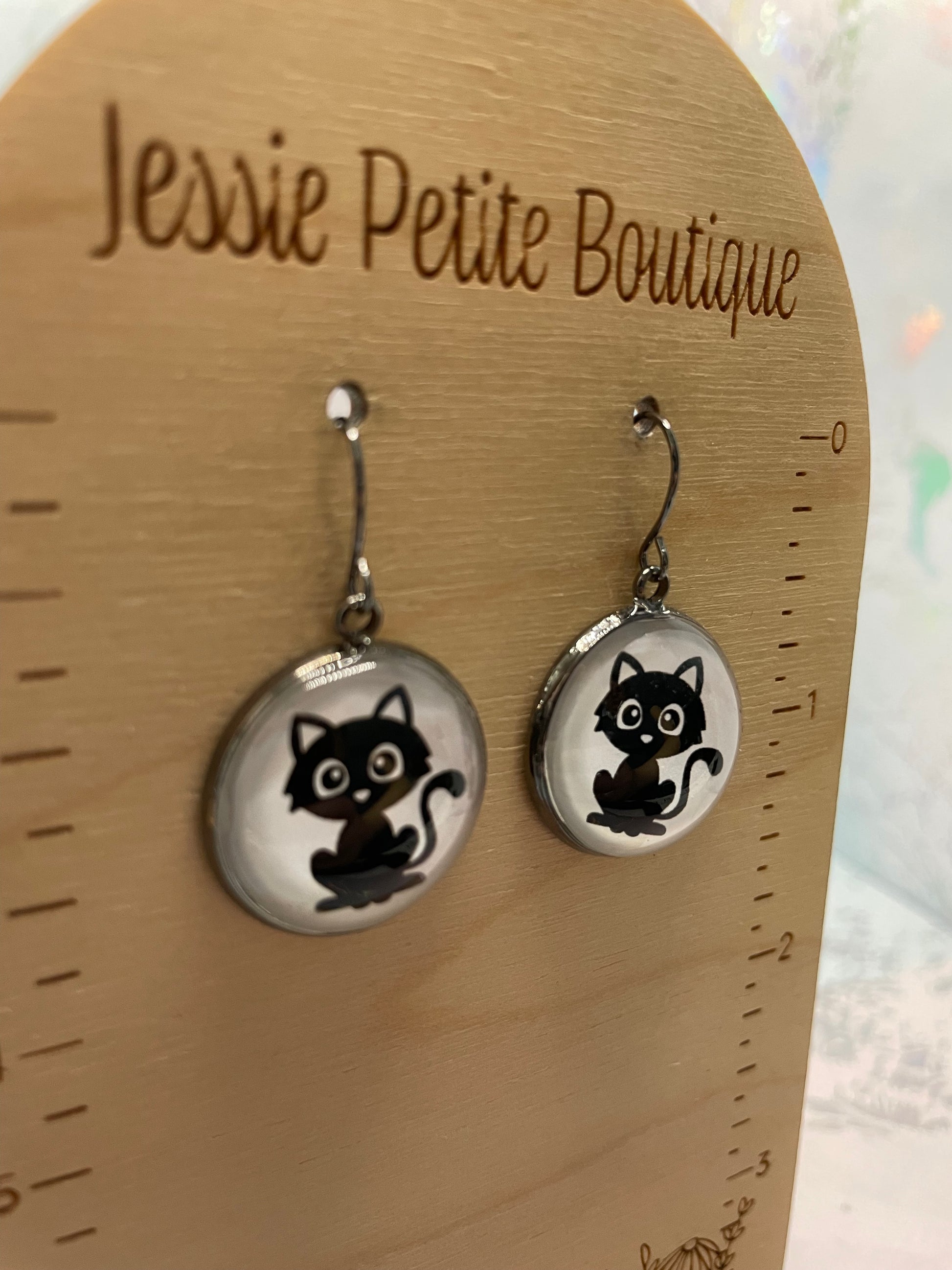 Cat-themed earrings on a wooden ruler with 'Jessie Petite Boutique' branding.