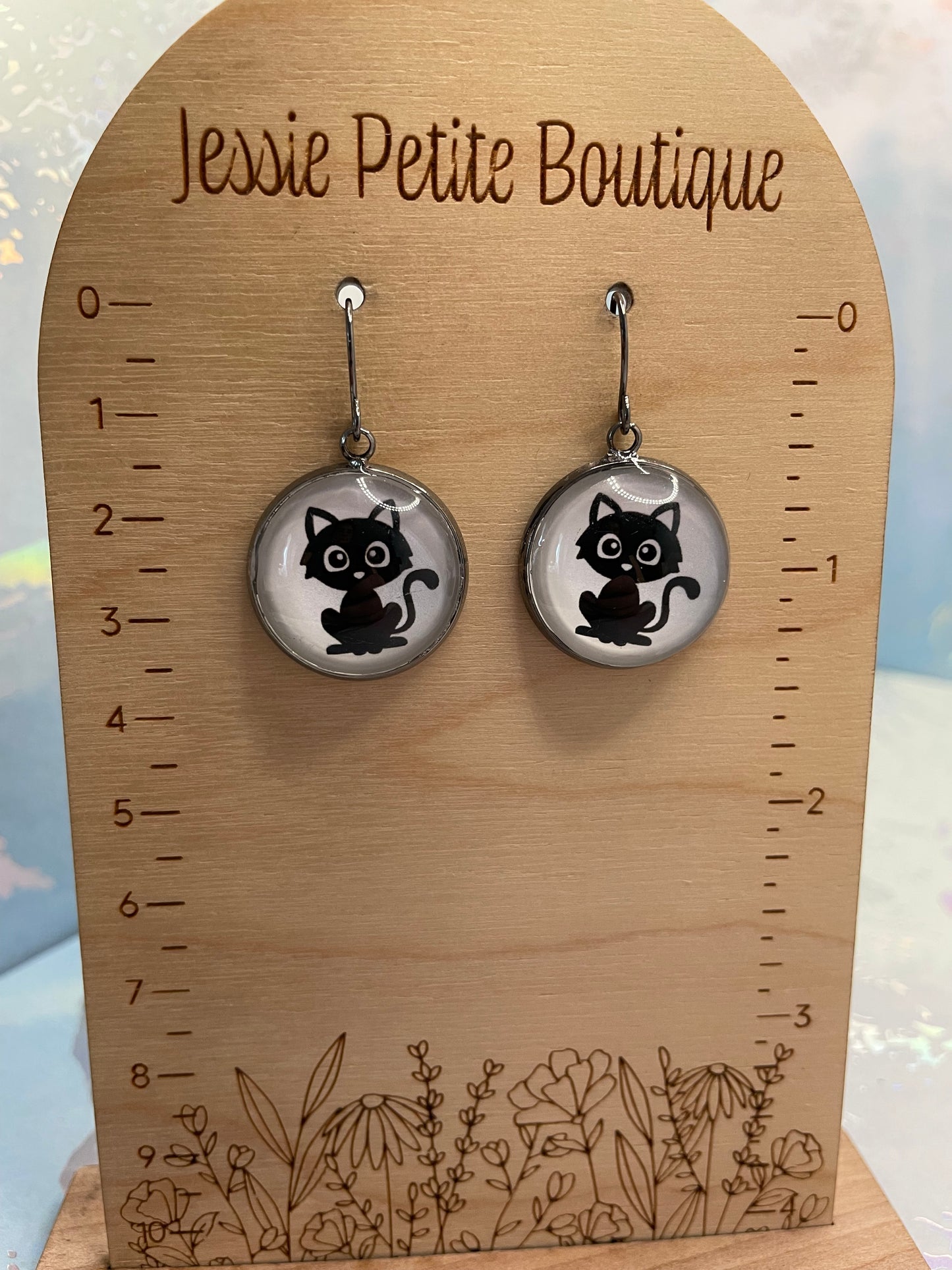Cat-themed earrings on a wooden card with measurements and floral designs, branded 'Jessie Petite Boutique'.