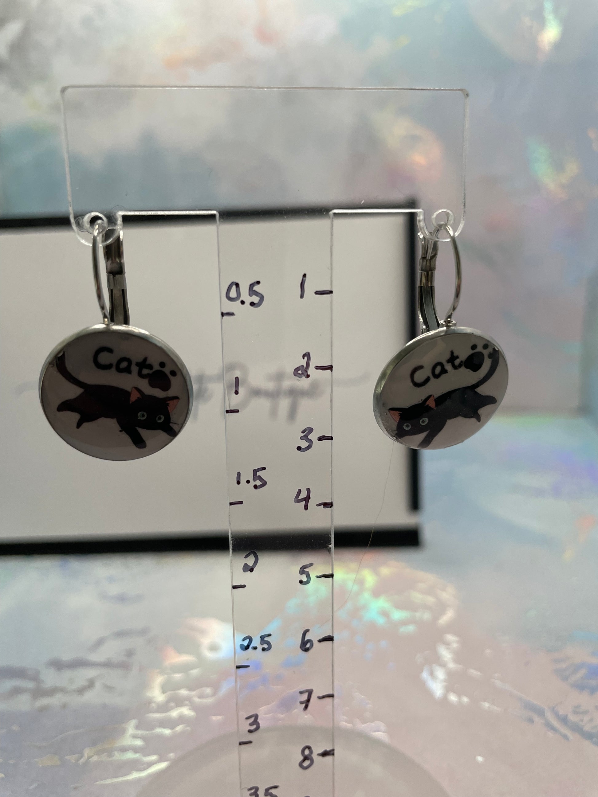 Pair of cat-themed earrings on a clear stand with measurement scale