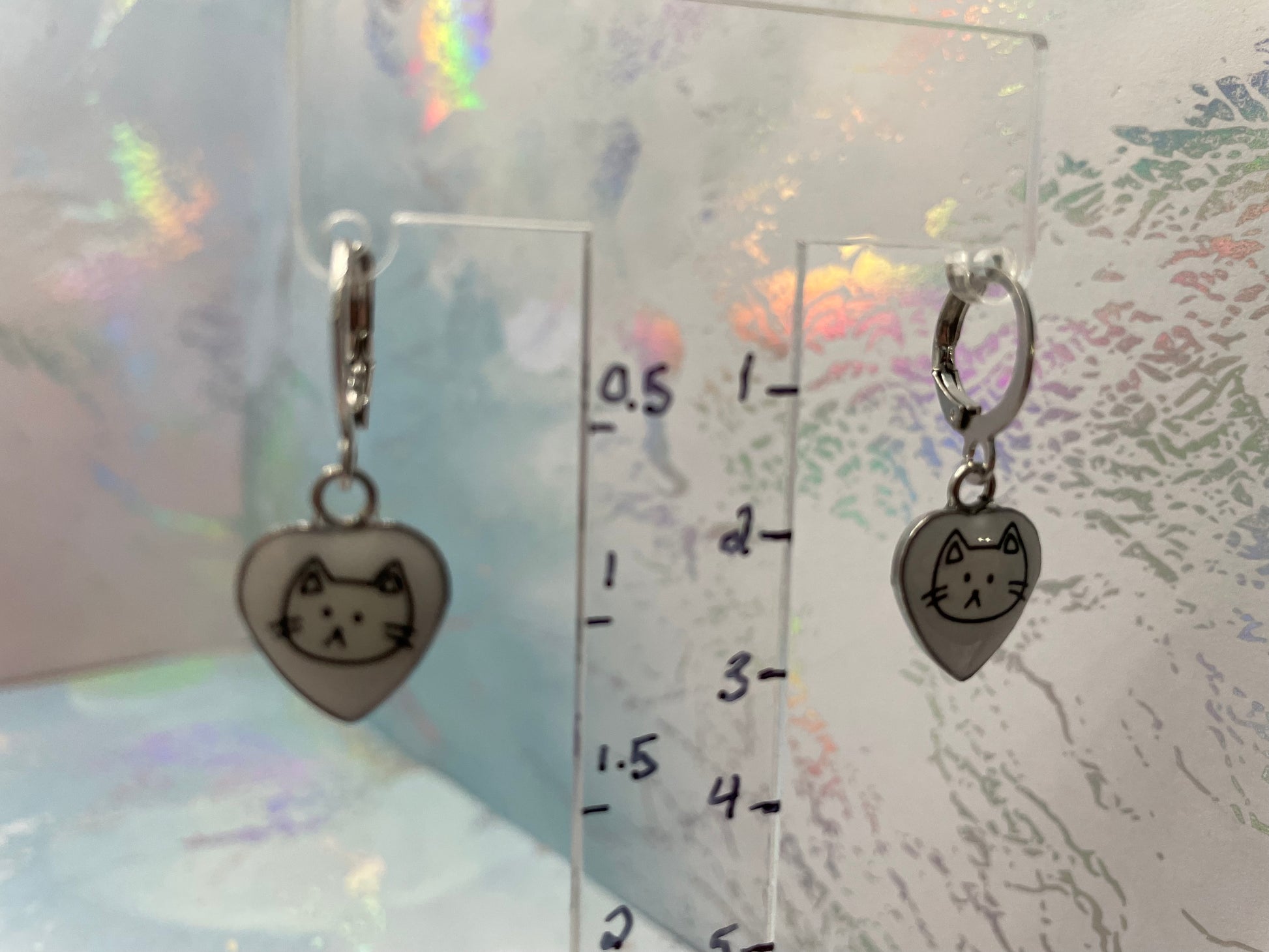 Heart-shaped earrings with cat design on a clear stand with measurement scale.