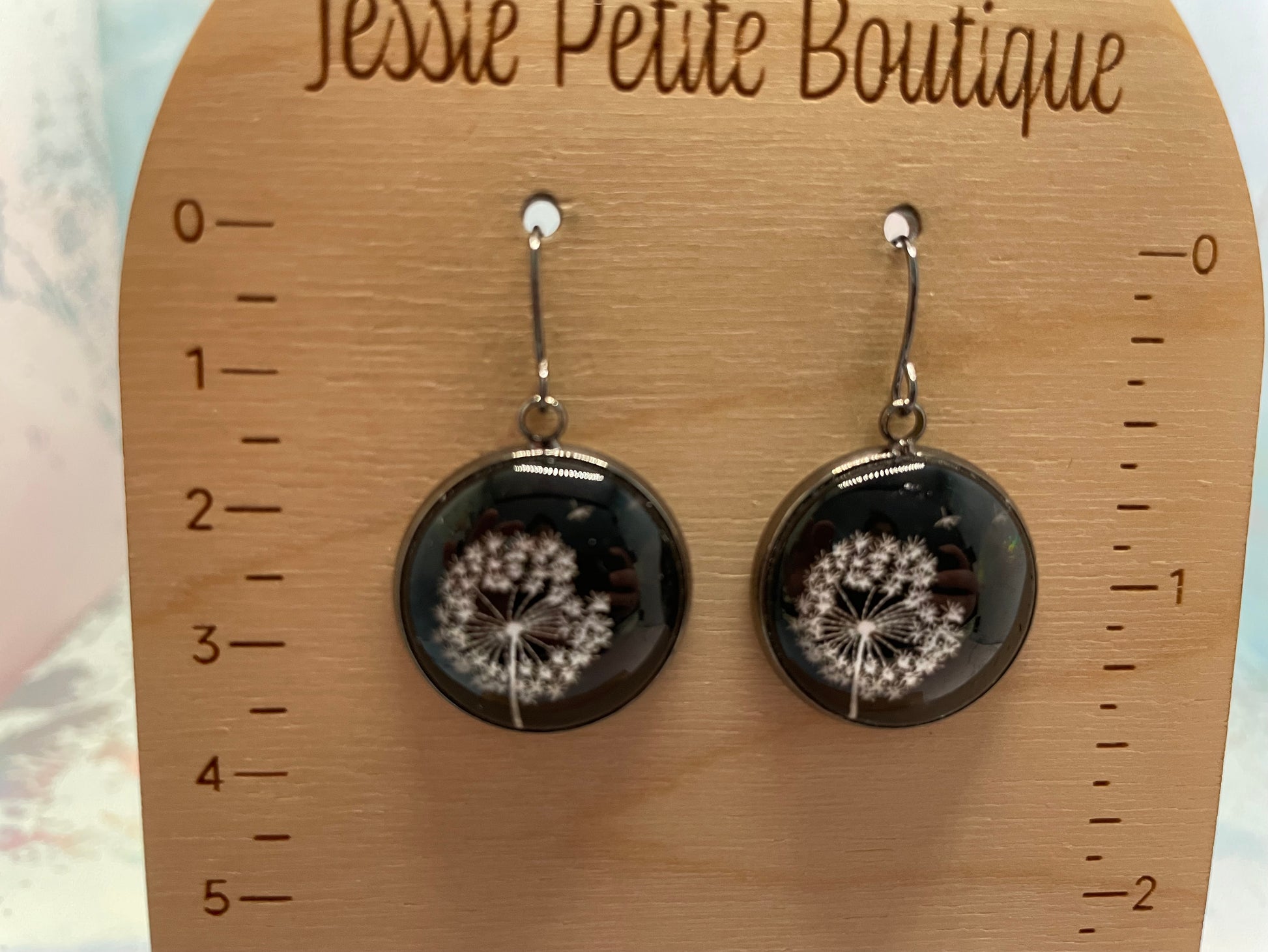 Dangle earrings with dandelion design on a wooden card with measurement scale, branded 'Jessie Petite Boutique'.