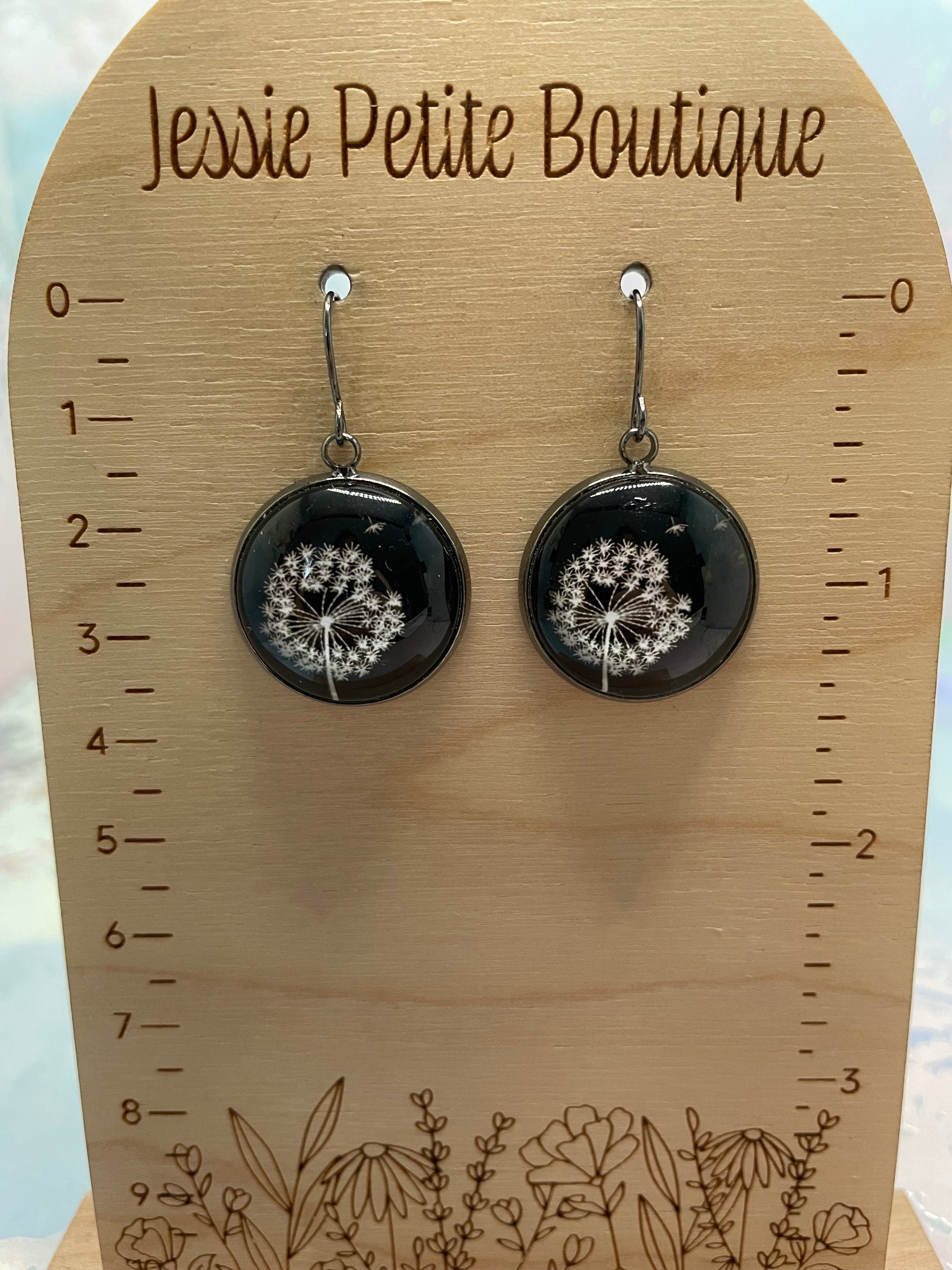 Black circular earrings with white floral design on a wooden card with measurements and floral patterns.