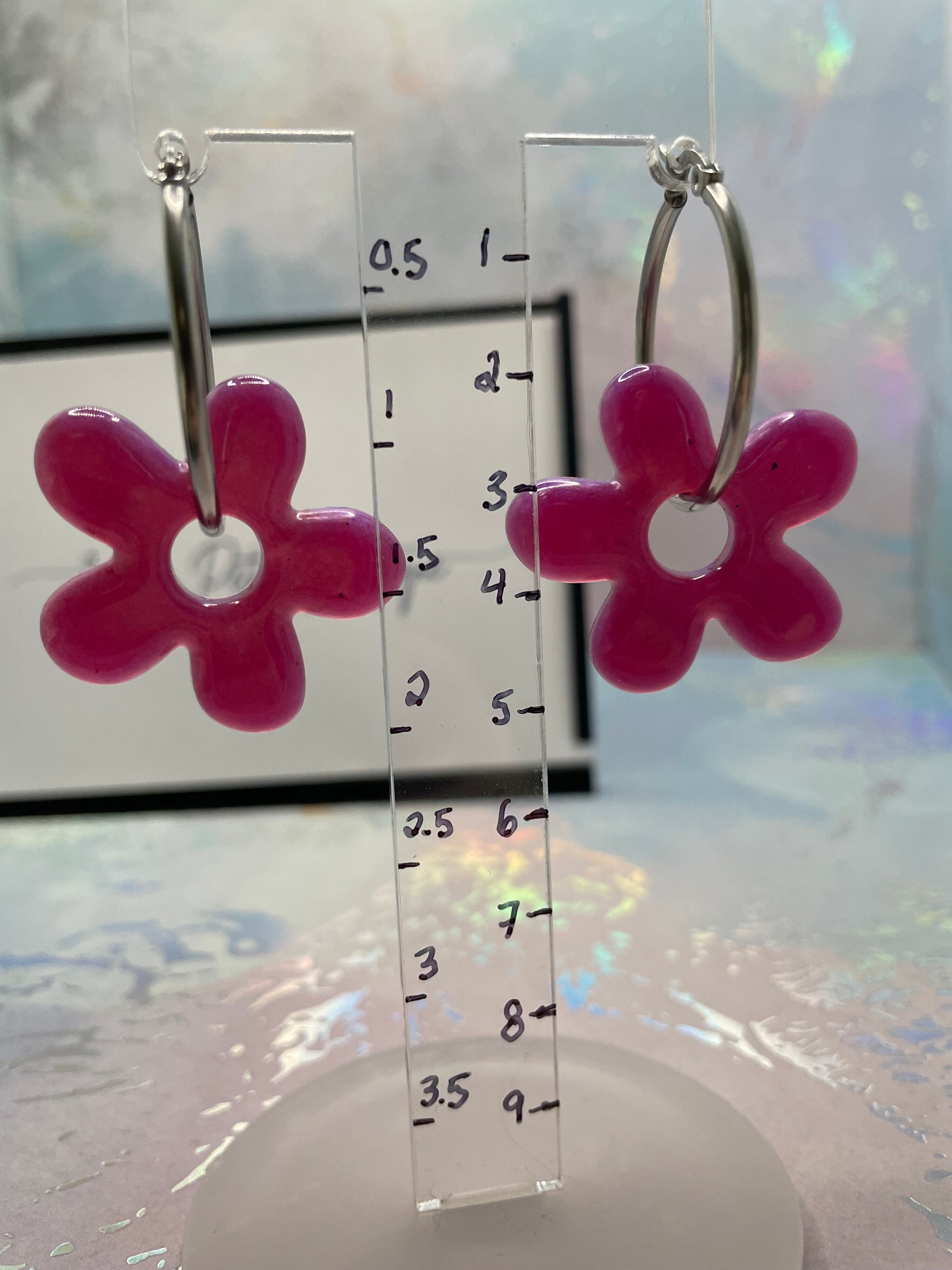 Pink flower-shaped earrings with a clear ruler for scale on a reflective surface.