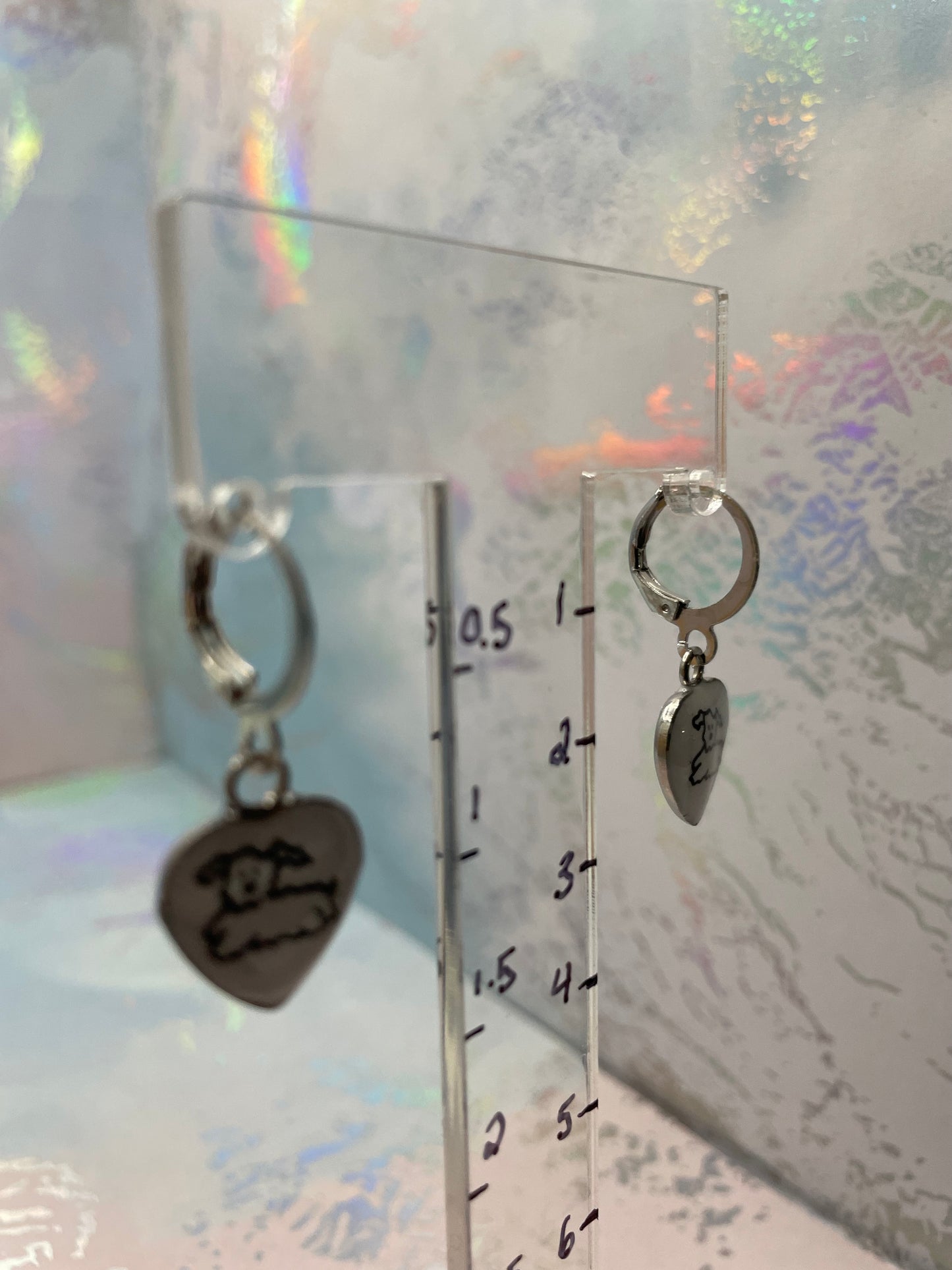 Silver earrings with heart-shaped charms on a clear stand against a reflective surface.