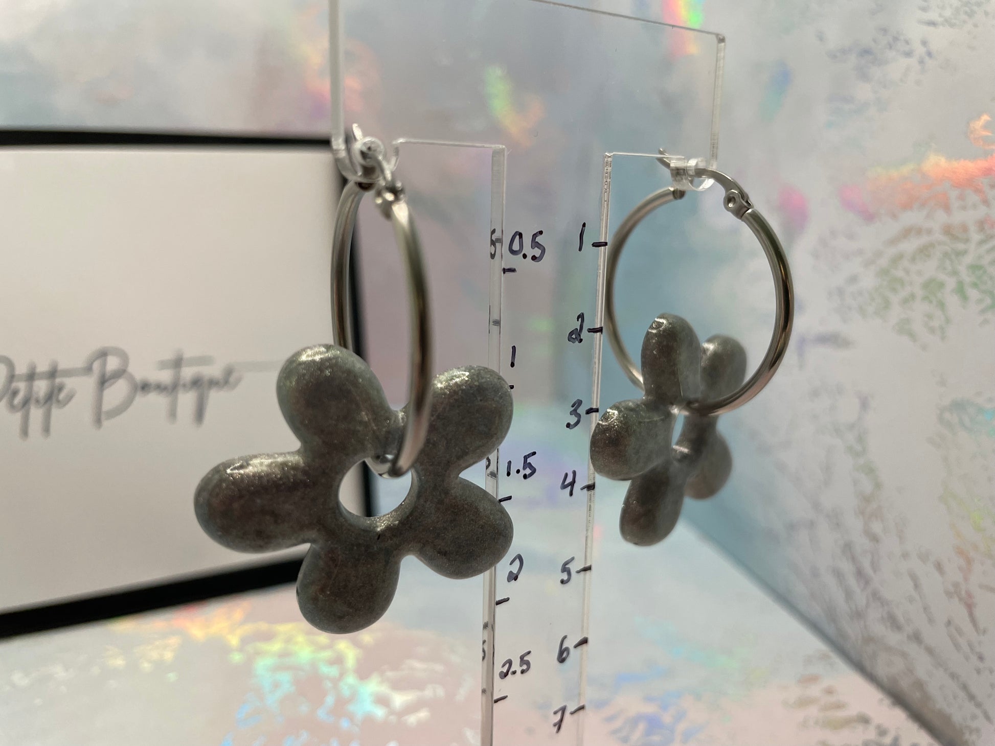 Flower-shaped earrings on a clear stand with measurement scale against a glittery background.