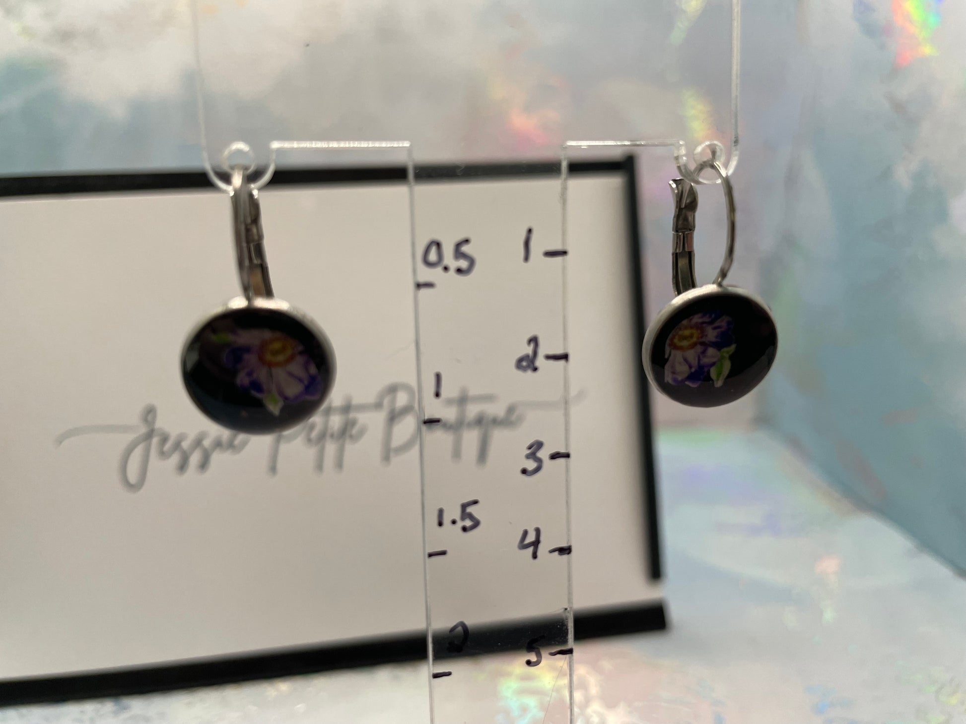 Pair of earrings on a clear stand with measurement scale, branded 'JessIe Petite Boutique'.