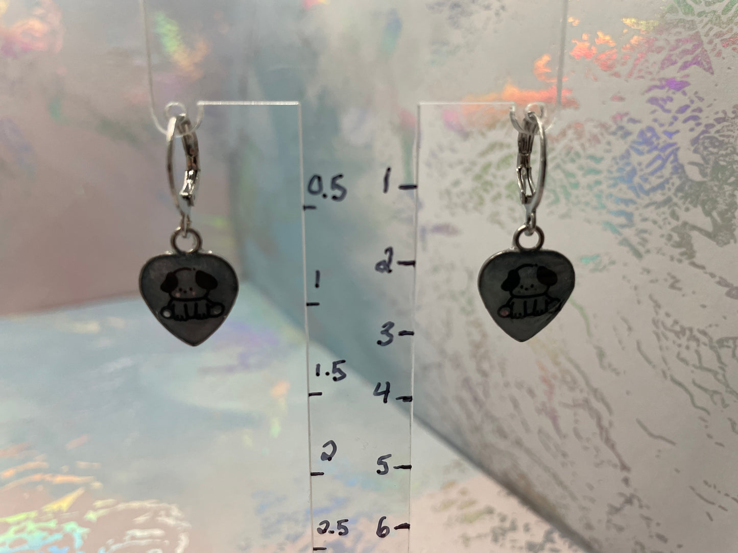 Heart-shaped earrings with a dog design on a clear ruler for scale against a textured background.