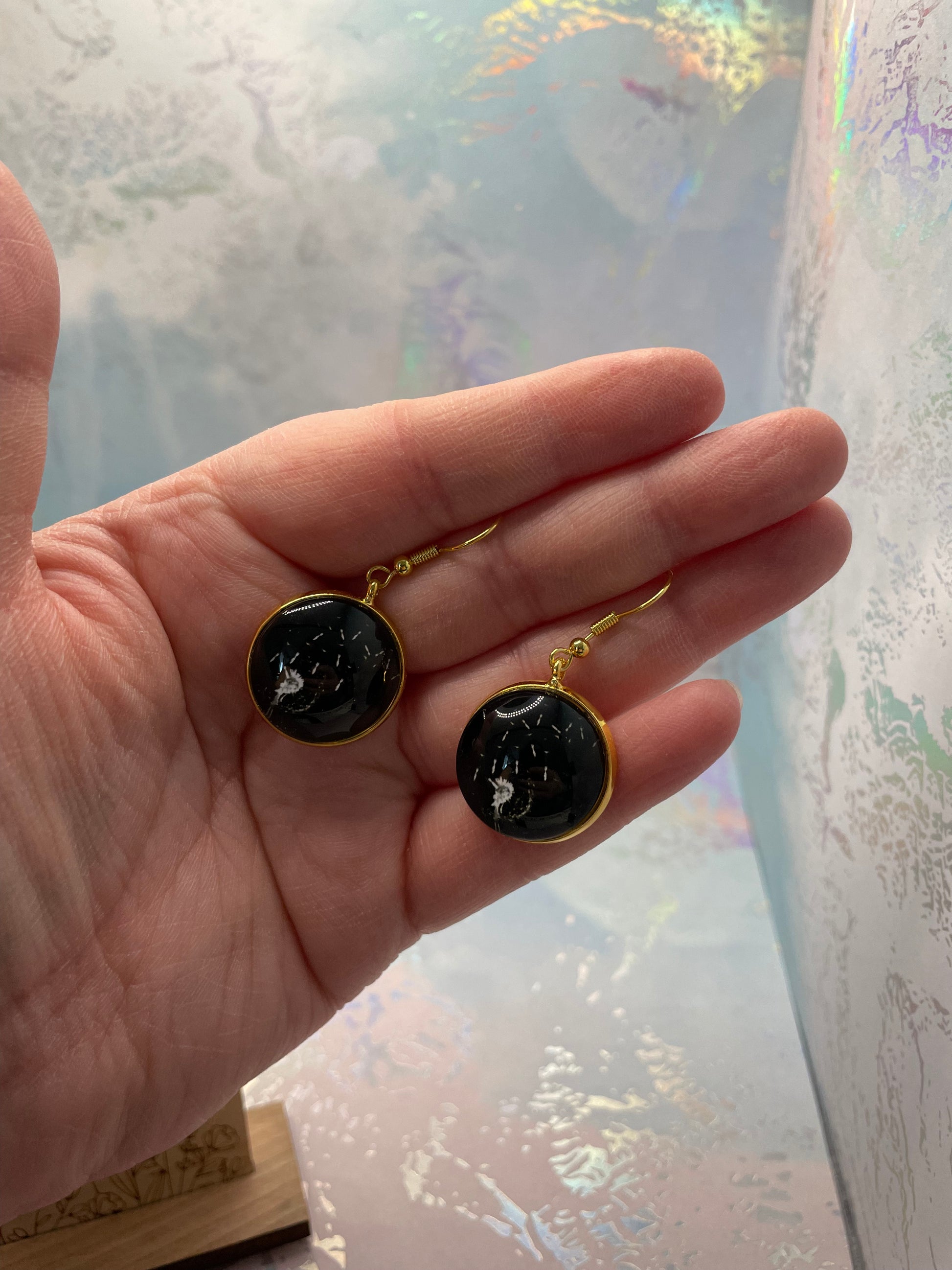 Hand holding a pair of Dandelion Round Cabochon Earrings with gold accents against a light background
