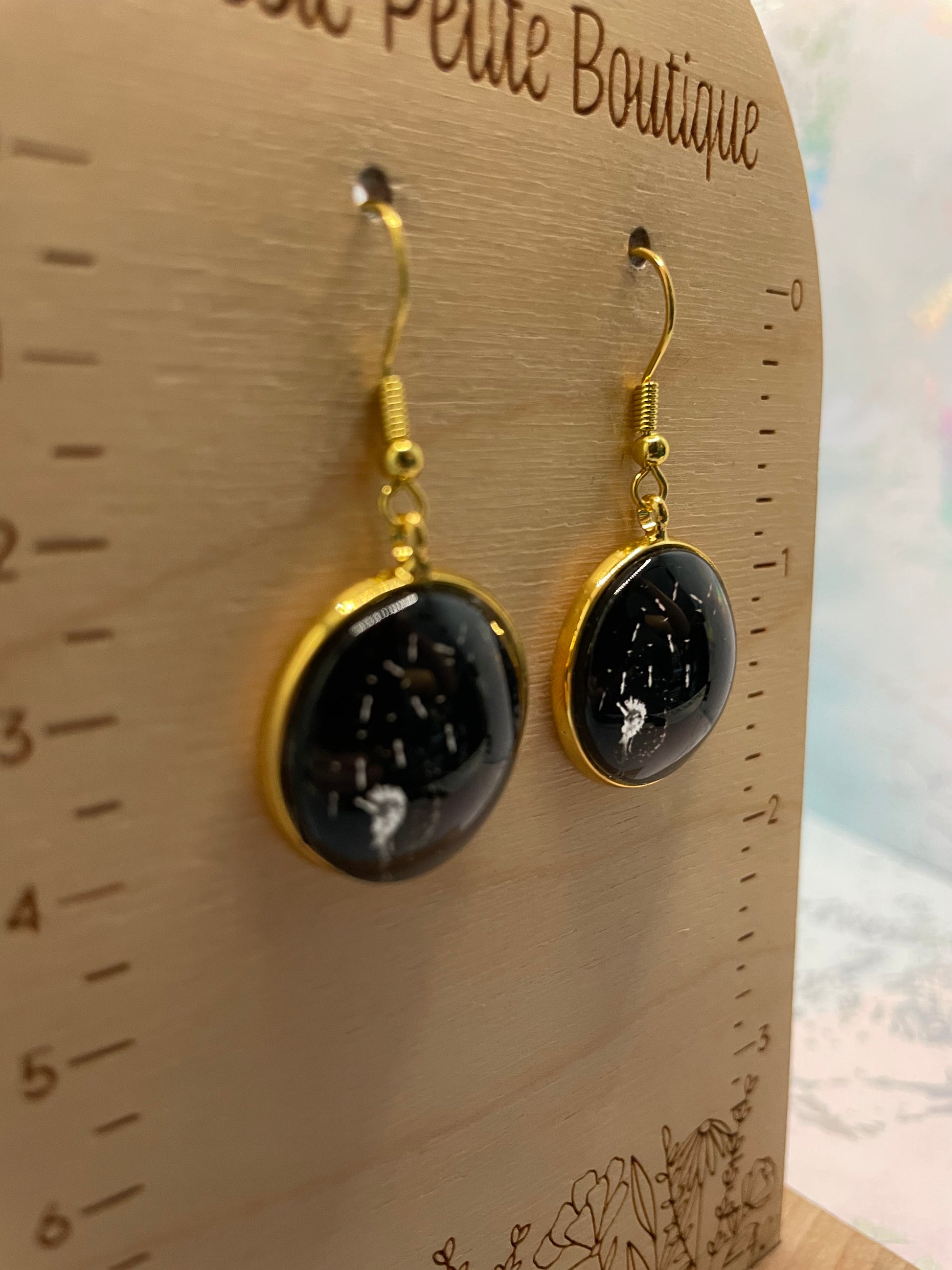 Pair of Dandelion Round Cabochon Earrings with gold accents on a wooden stand with measurement scale.