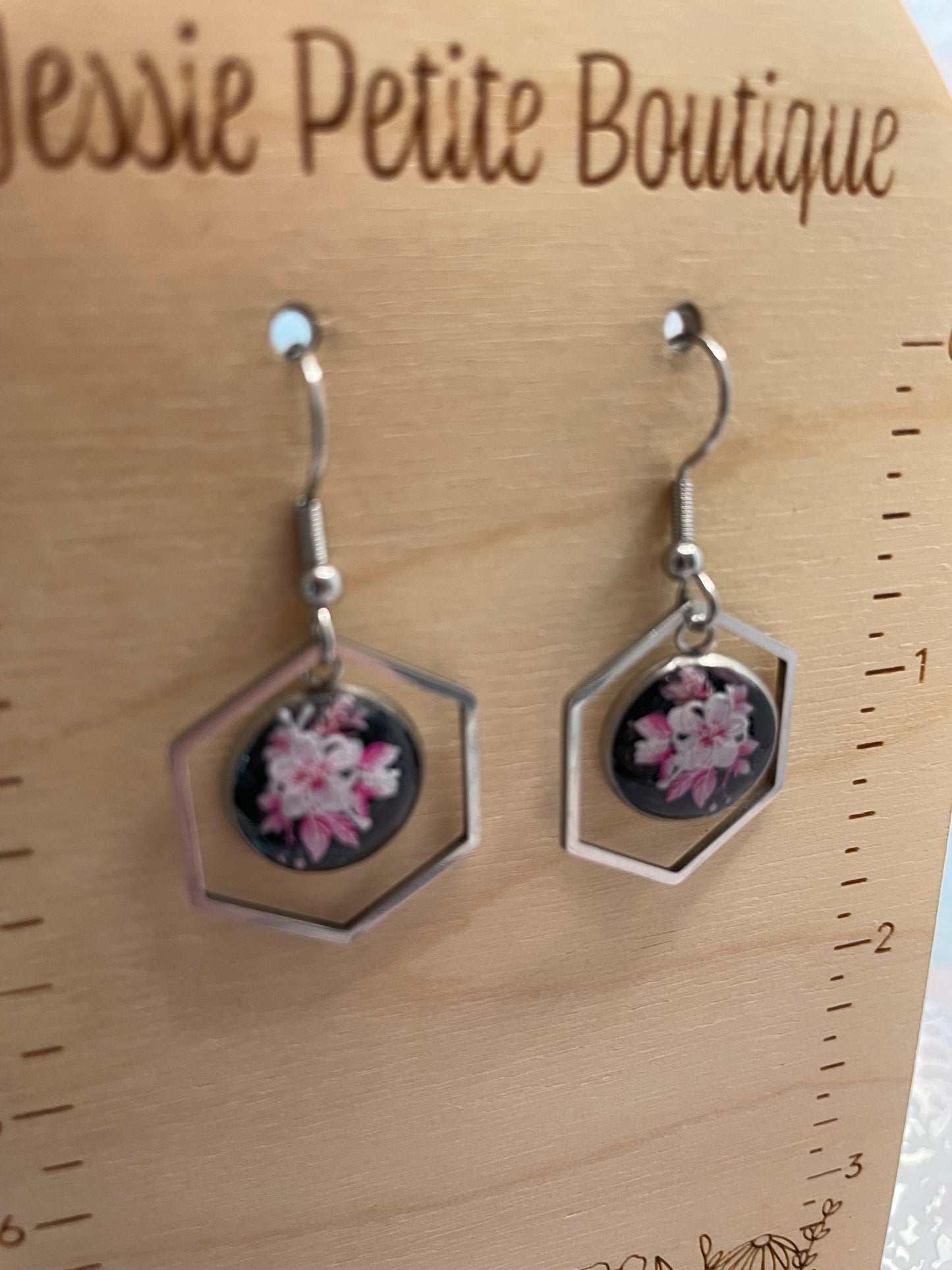 Hexagonal earrings with floral design on a wooden surface with 'Jessie Petite Boutique' branding.
