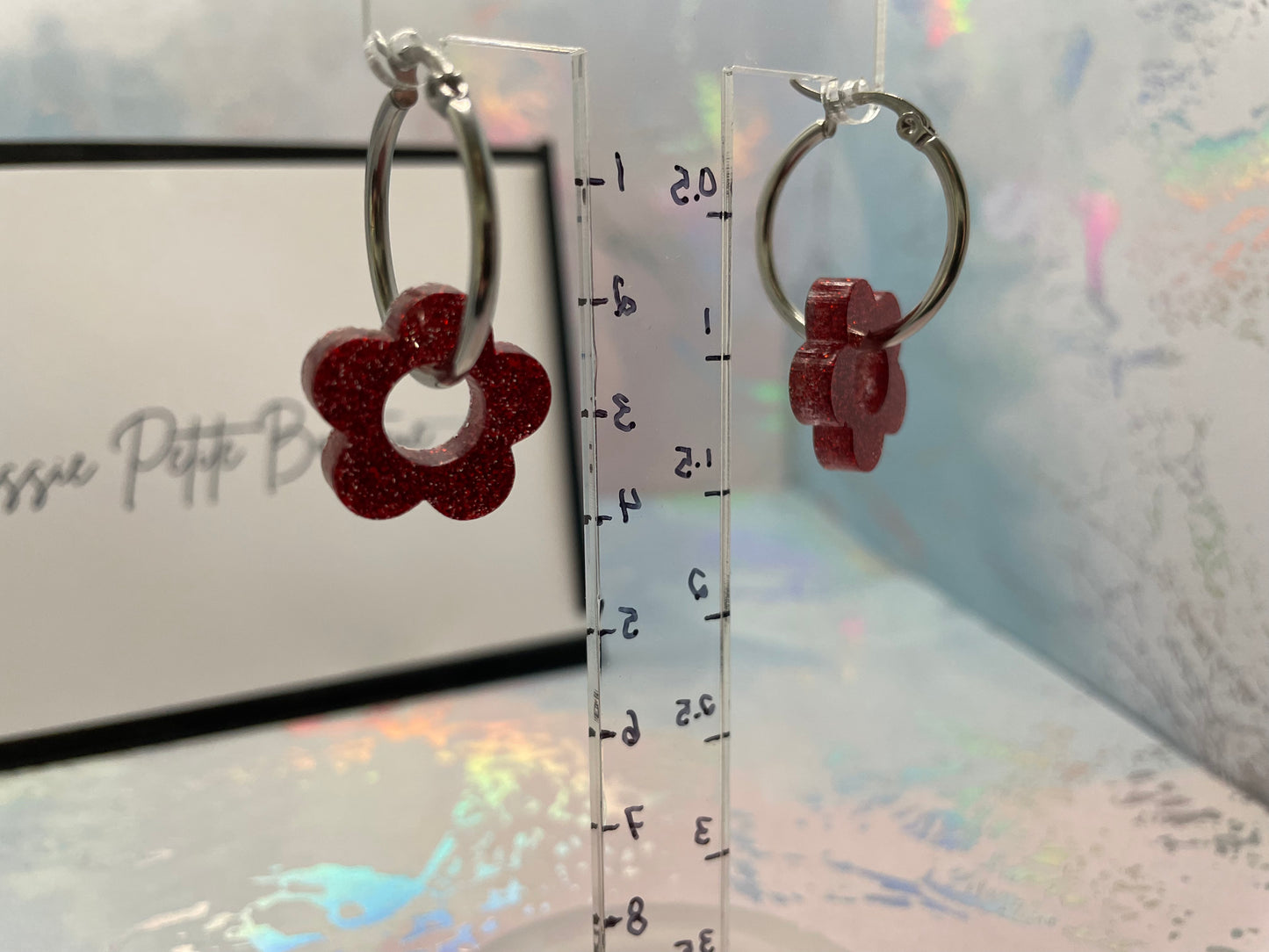 Silver hoop earrings with red flower-shaped charms on a clear ruler for scale.