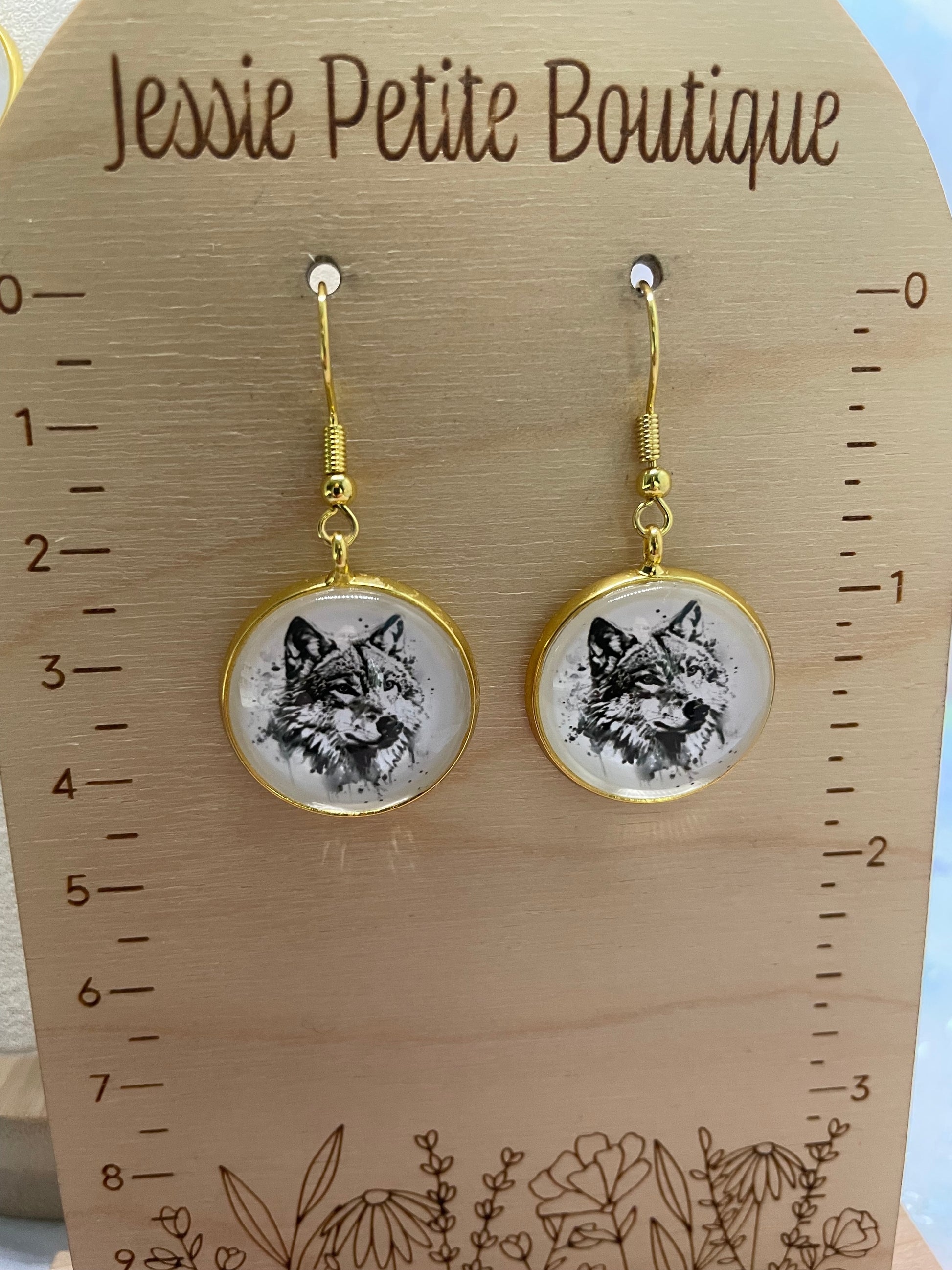 Pair of earrings with wolf design on a wooden card with measurements, branded 'Jessie Petite Boutique'.