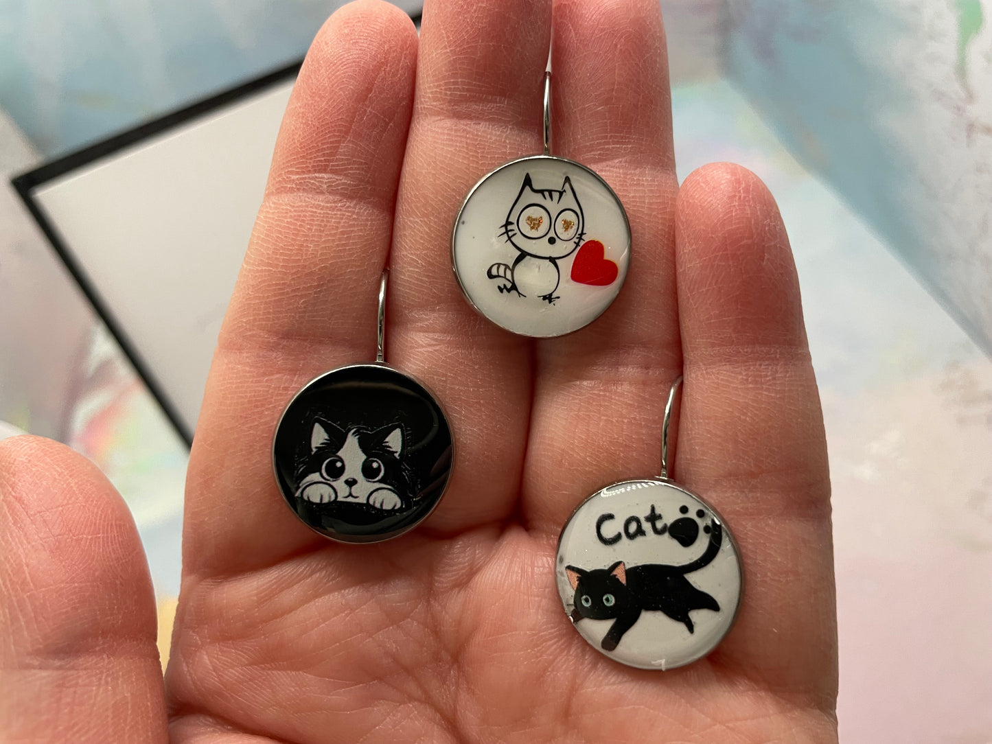 Three cat-themed earrings held in a hand with a blurred background