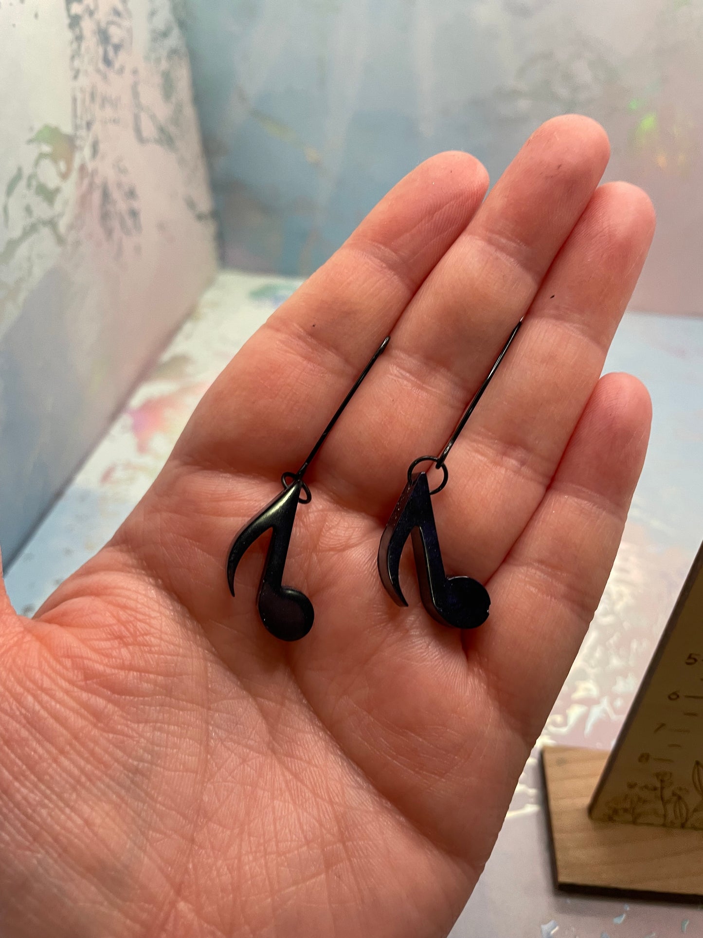 Hand holding two Dark Blue Music Note Earrings a blurred indoor background