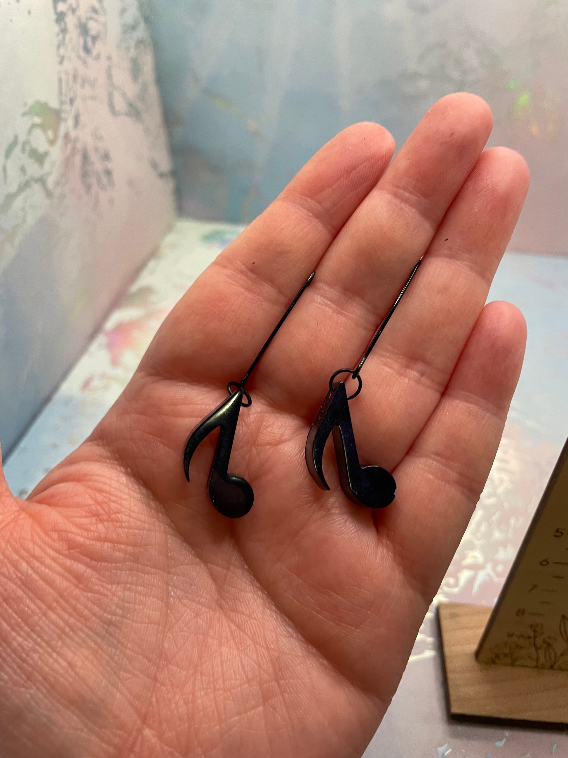 Hand holding two Dark Blue Music Note Earrings a blurred indoor background