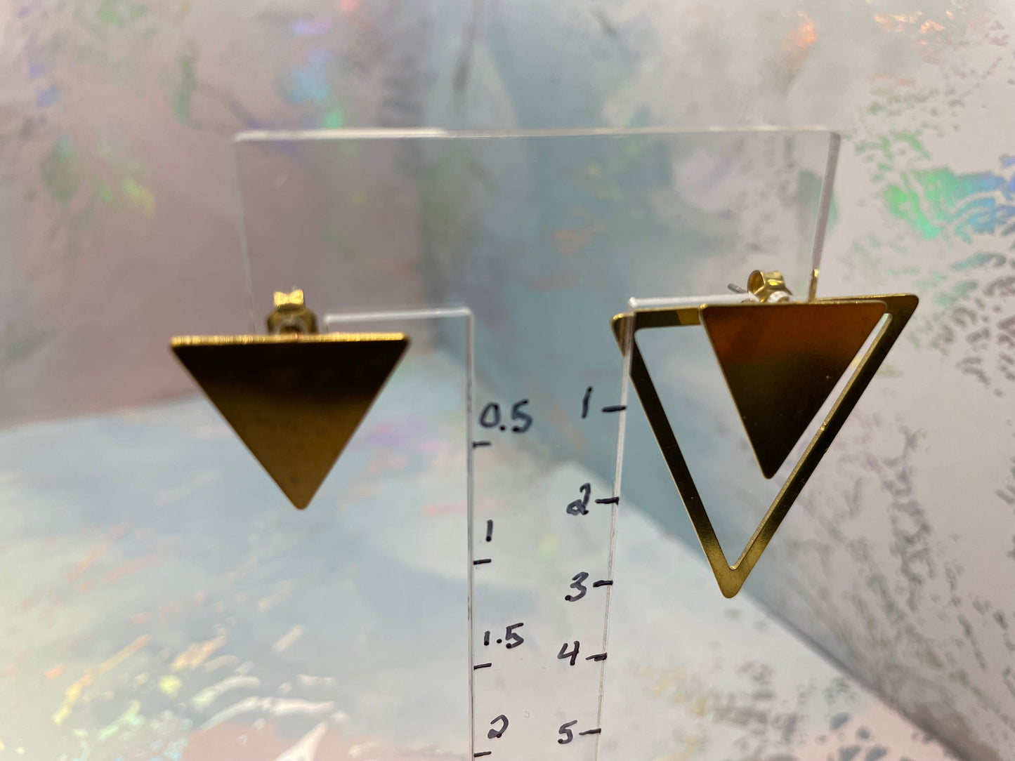 Gold triangle-shaped earrings on a clear stand with measurement scale against a patterned background
