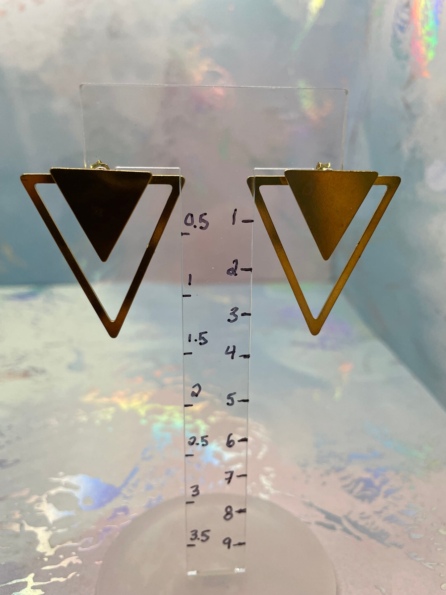 Gold triangle earrings with a clear ruler for scale on a reflective surface.