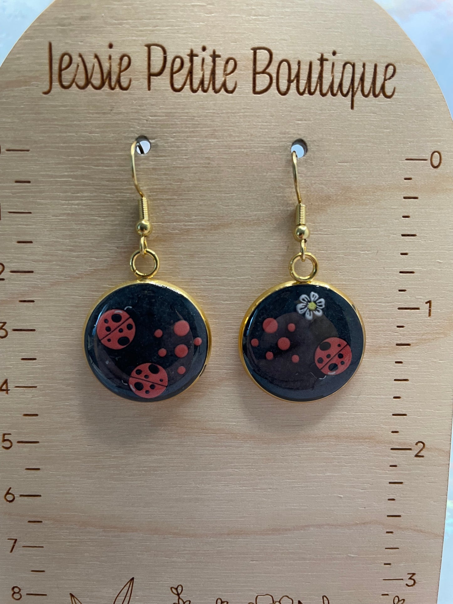 Black earrings with red polka dots+ladybugs on a wooden surface with 'Jessie Petite Boutique' branding.
