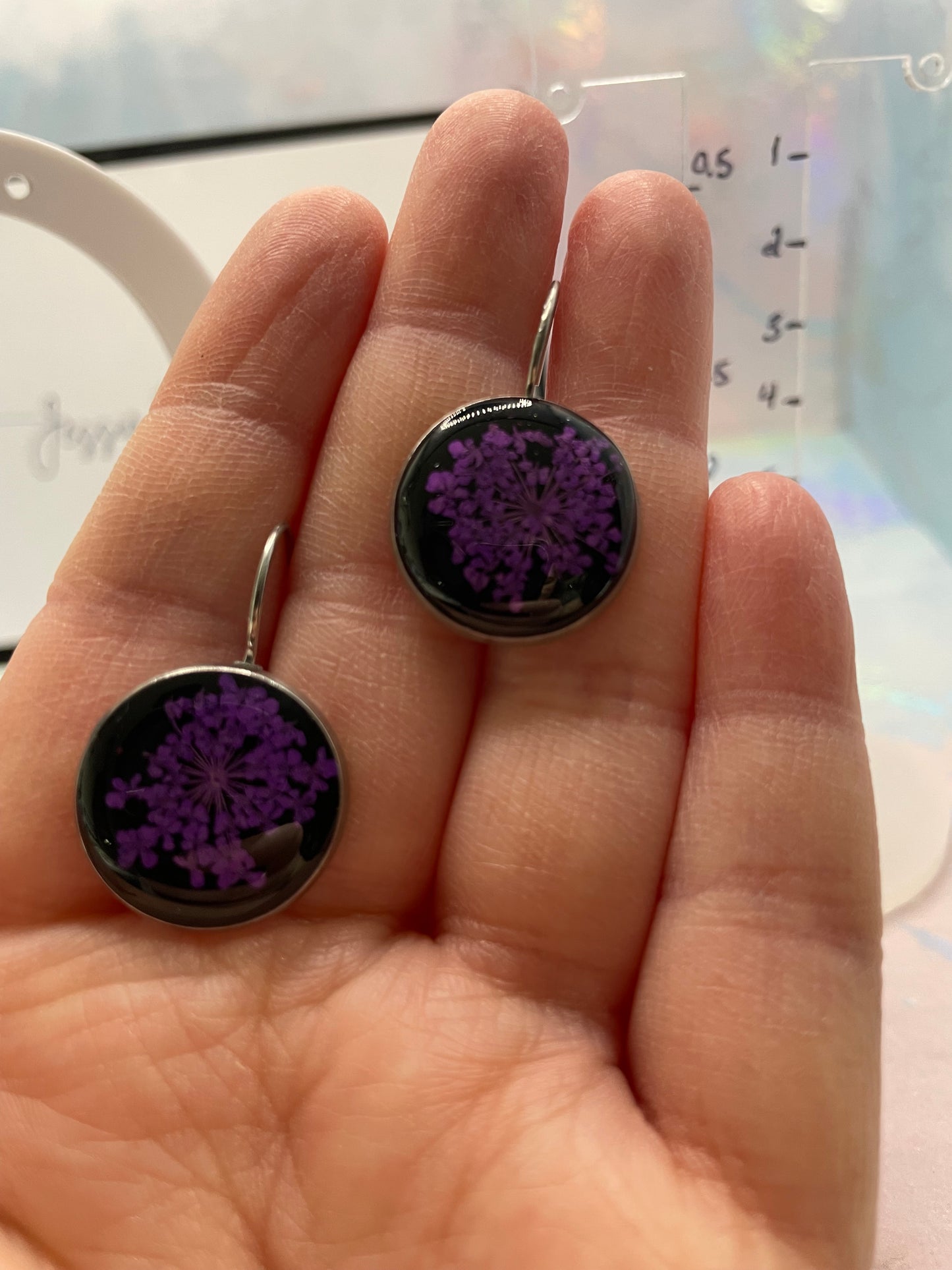 Purple Queen Anne’s Lace on Black Bezel Earrings held between fingers with a blurred background
