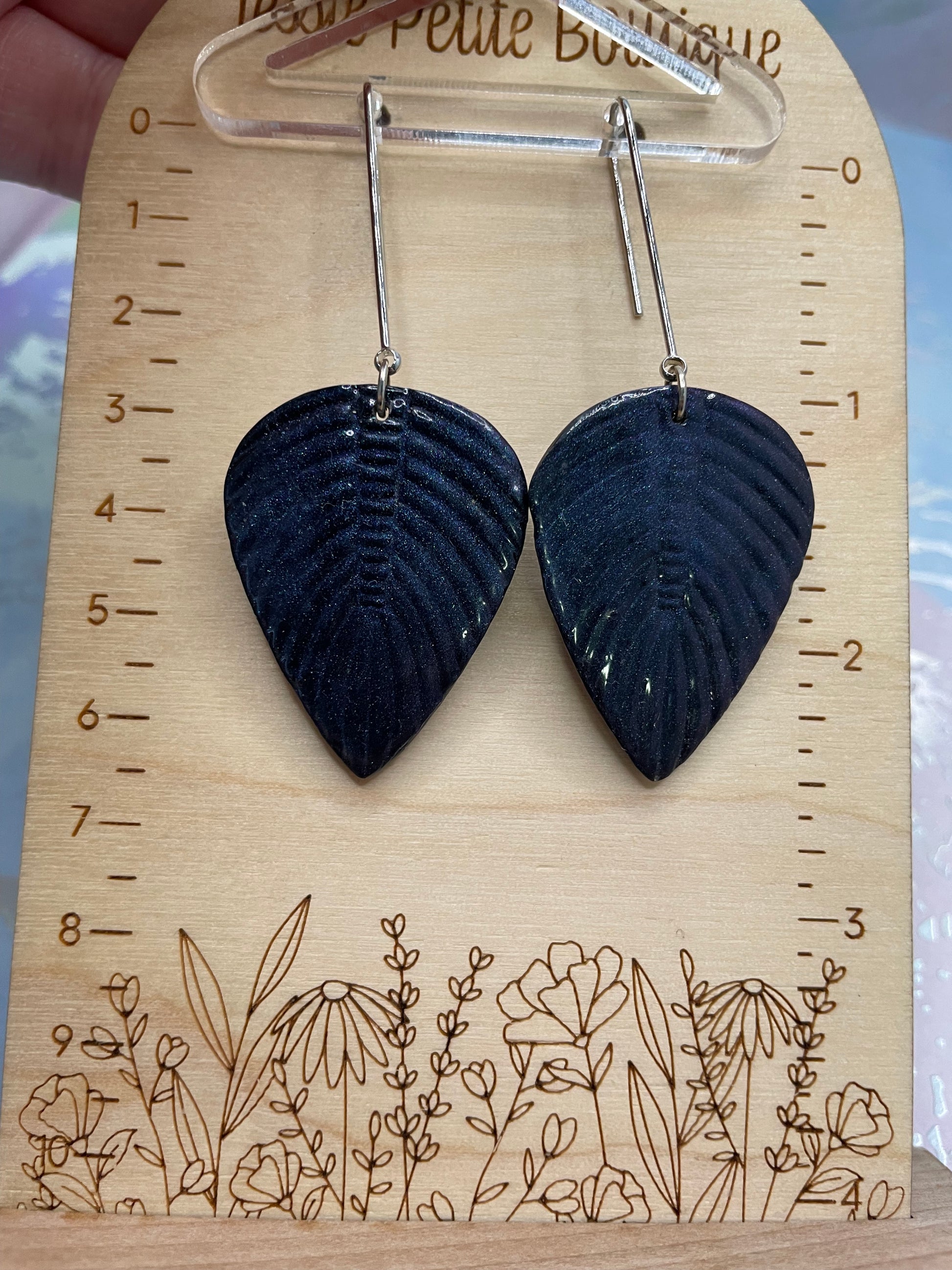 Black leaf-shaped earrings on a wooden display with floral designs and measurements.