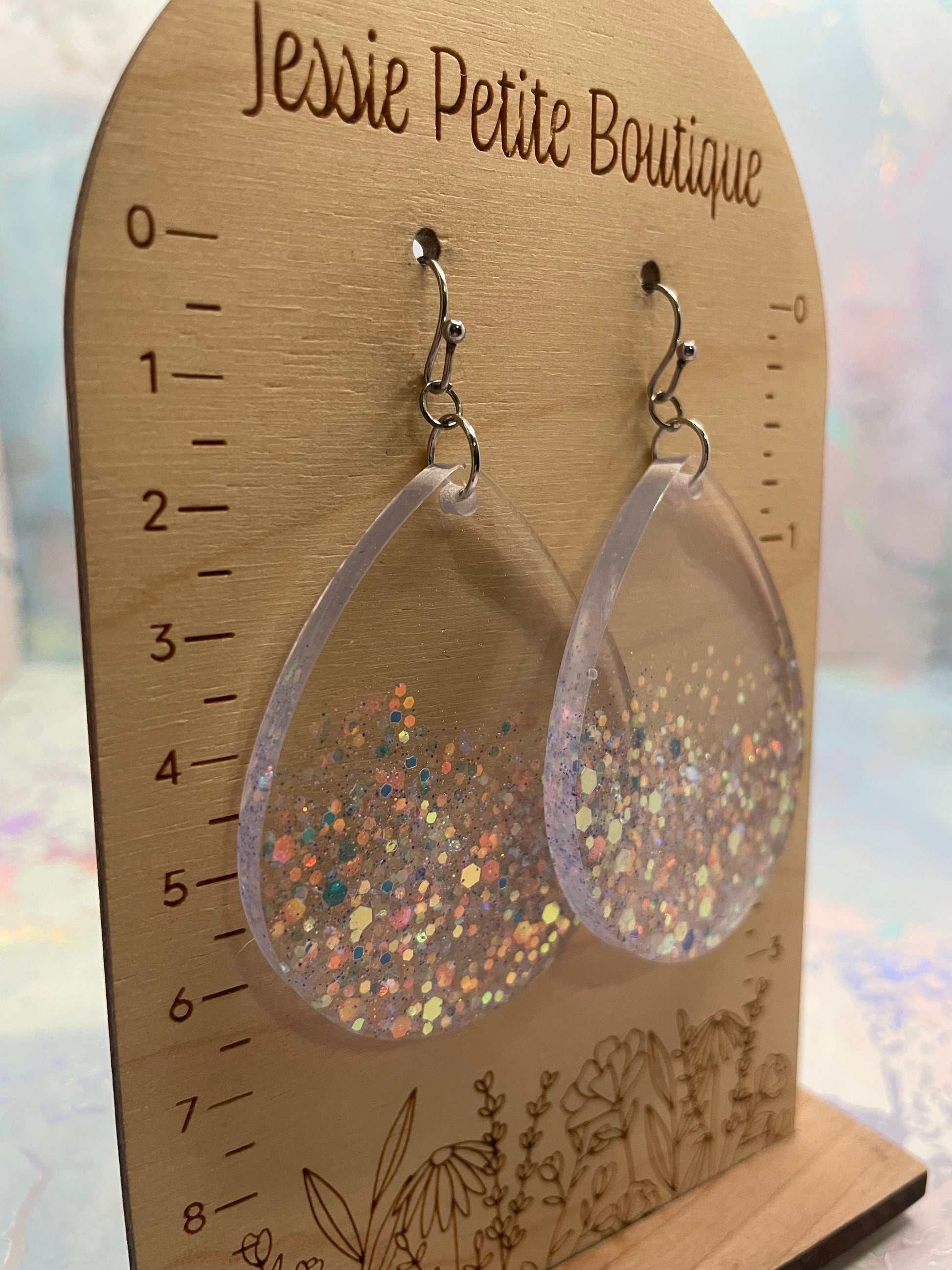 Clear resin earrings with glittery interior on a wooden card with measurements, branded 'Jessie Petite Boutique'.