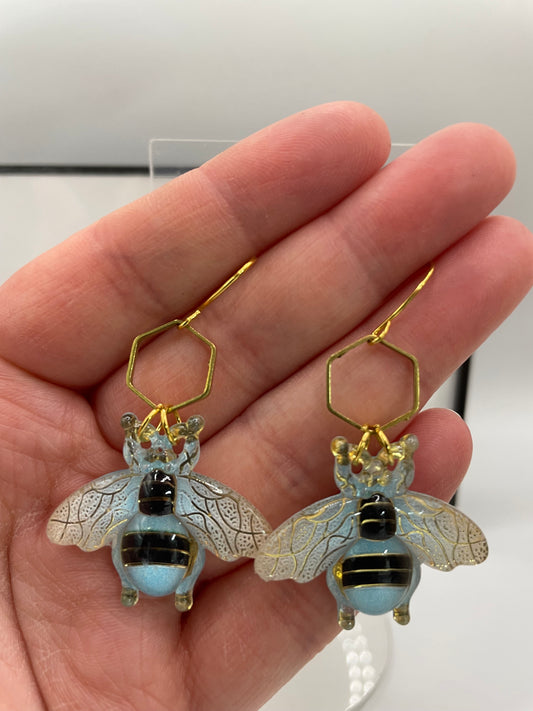 Hand holding bee-shaped earrings with gold hexagonal hooks.