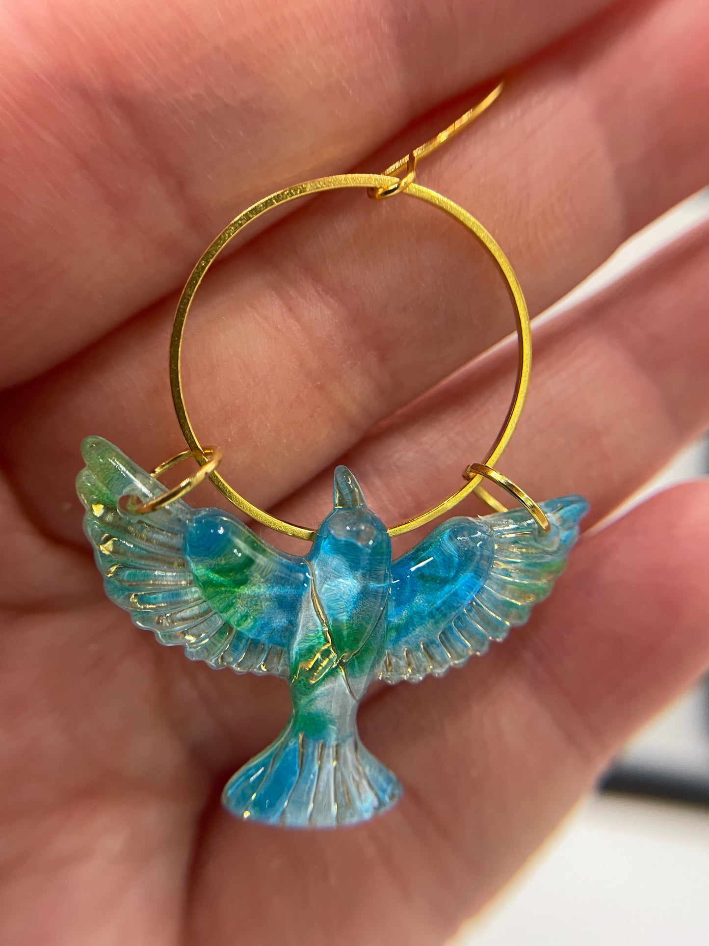 Gold hoop earring with blue and green bird design held in a hand.