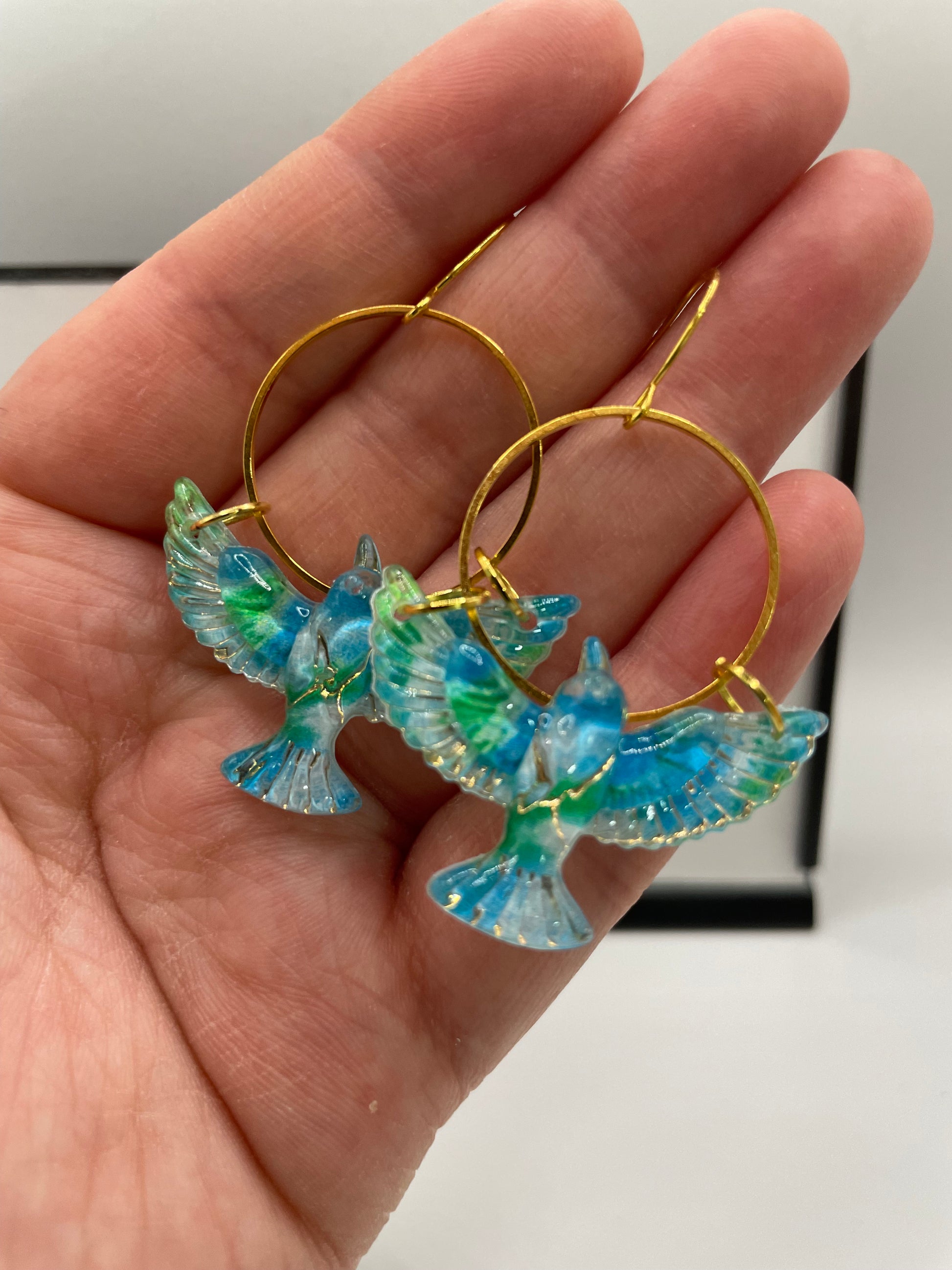 Hand holding gold hoop earrings with blue and green floral designs.