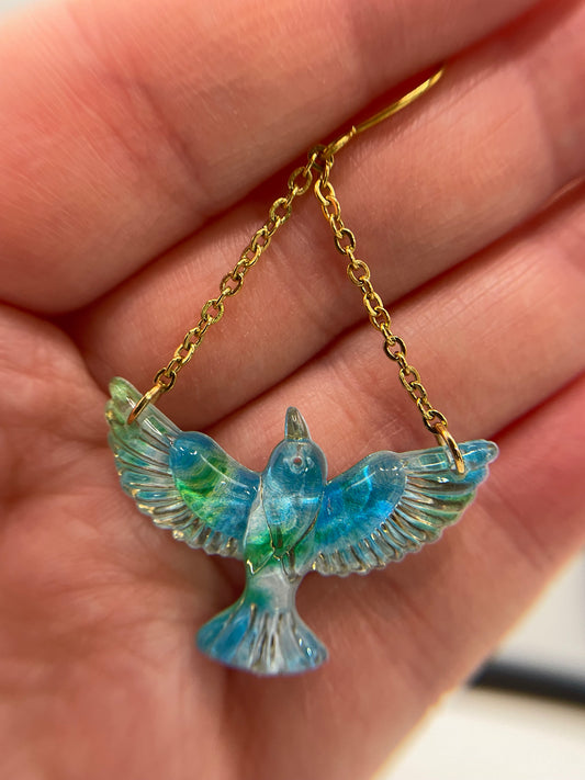 Blue bird-shaped pendant on a gold chain held in a hand.
