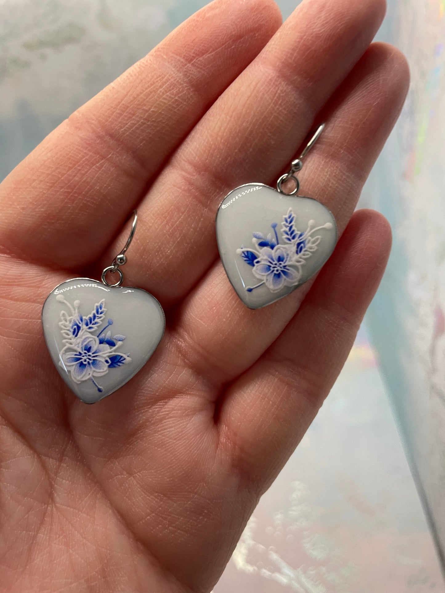 Heart-shaped earrings with blue floral designs held in a hand against a light background