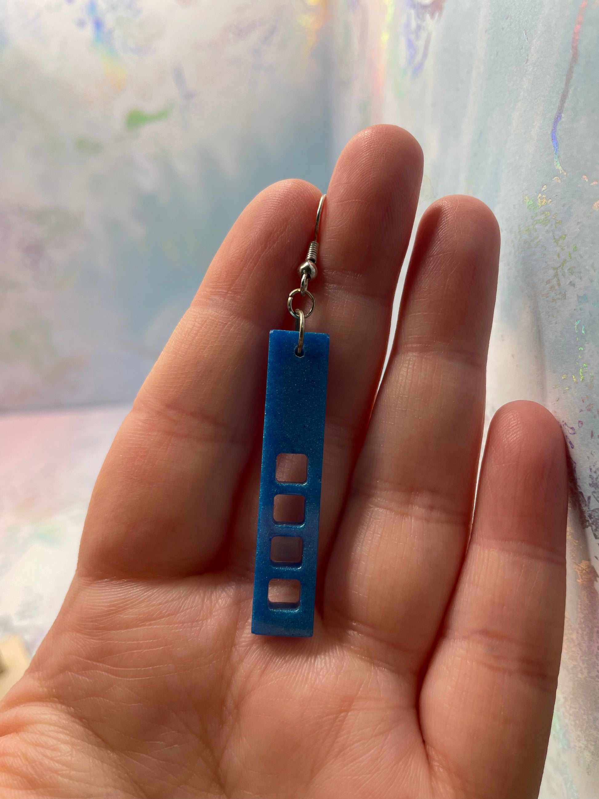Hand holding a blue rectangular earrings with cut-out sections against a blurred background
