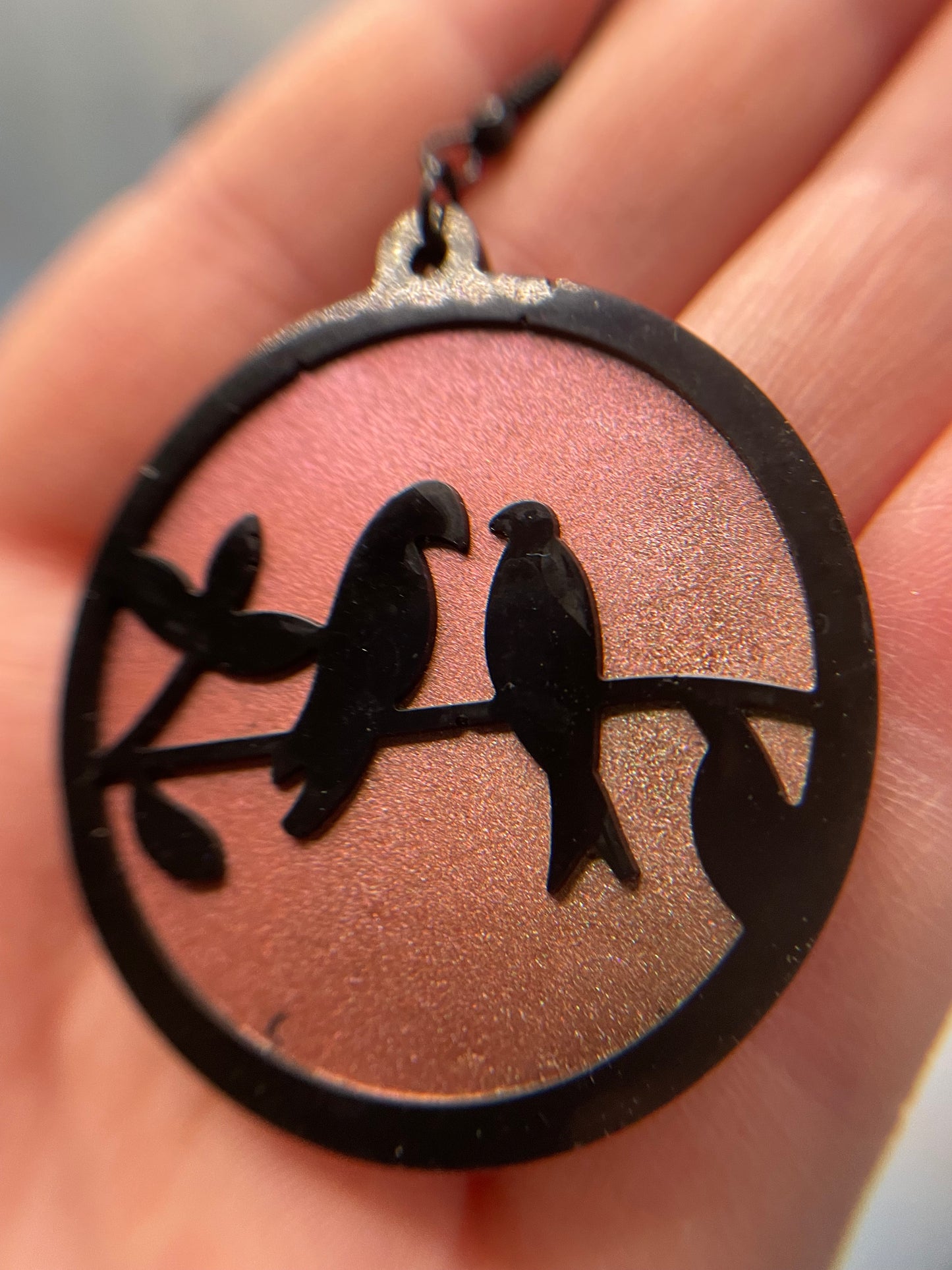 A hand holding a round earrings featuring a black love bird silhouette on a chameleon-like textured background.