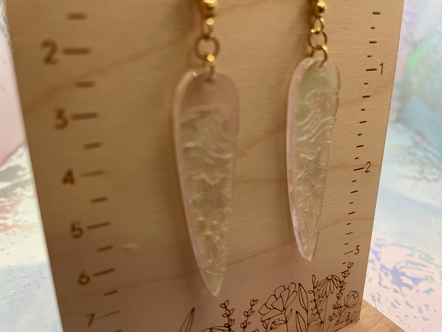 Pair of long, thin earrings on a wooden stand with measurement markings.