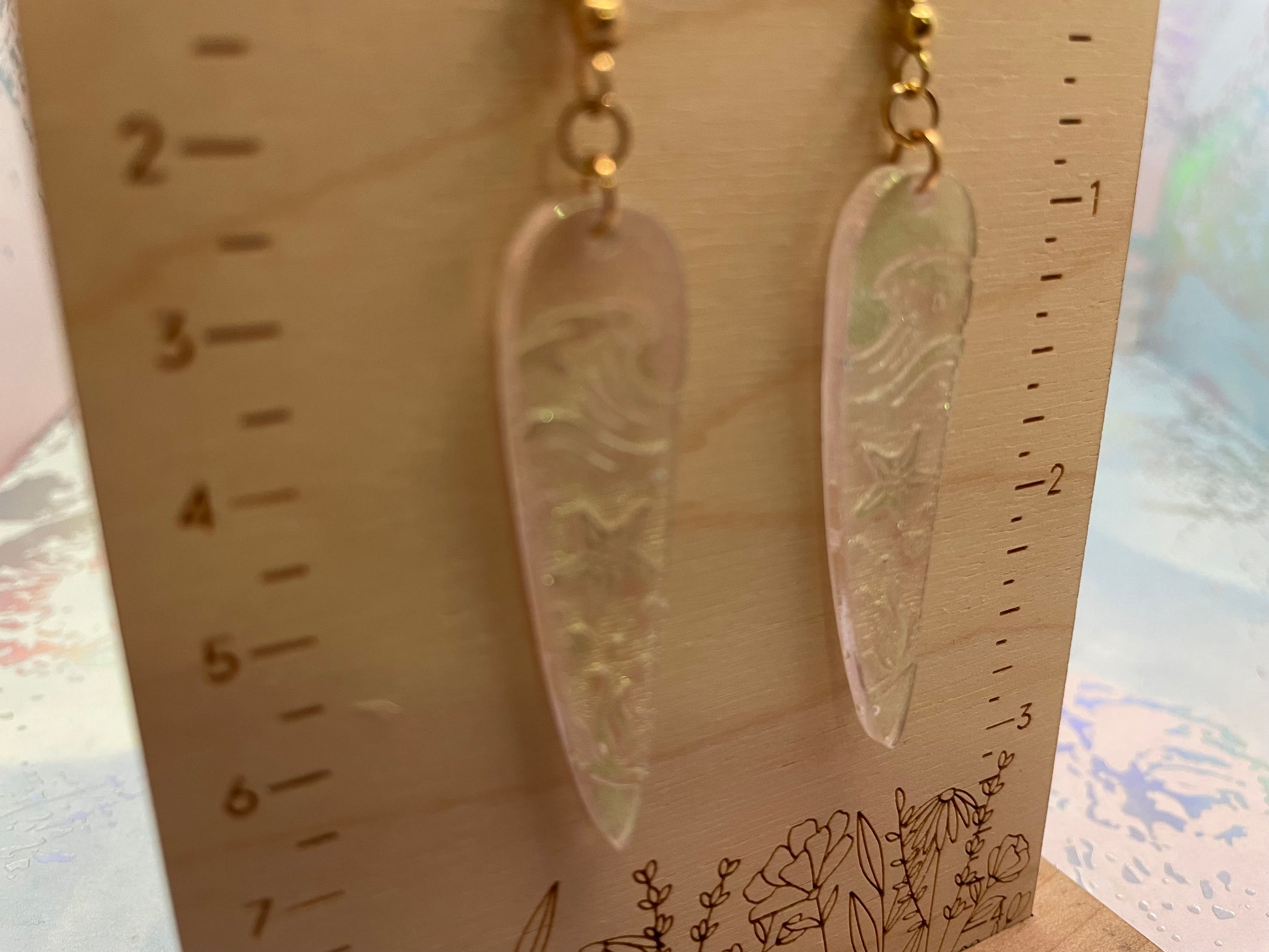 Pair of long, thin earrings on a wooden stand with measurement markings.