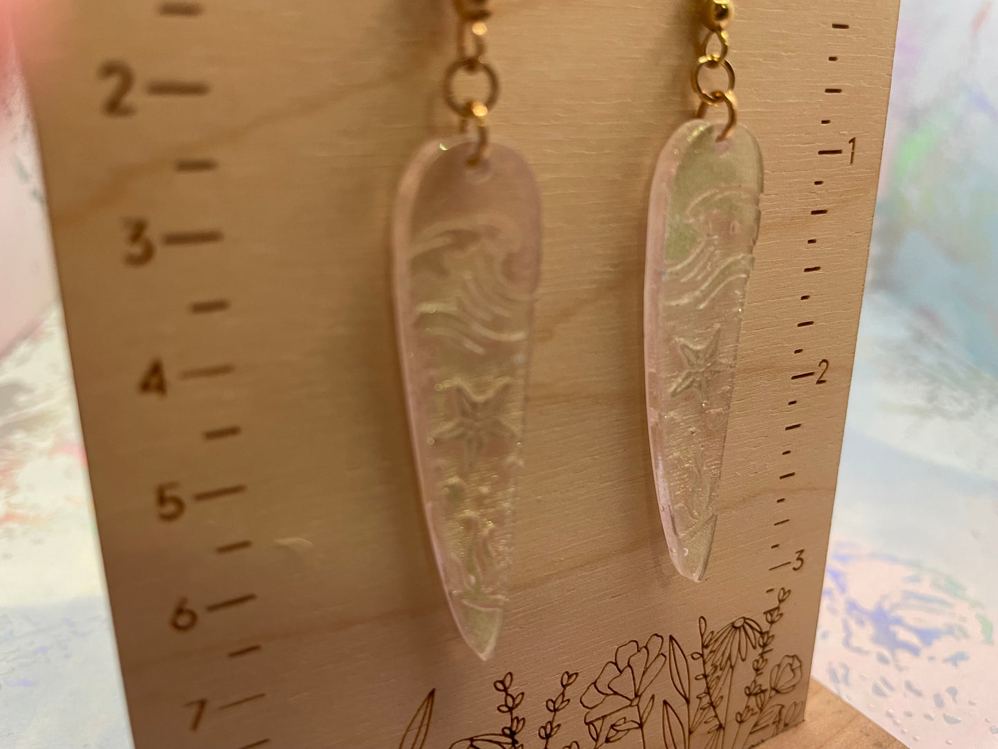 Pair of long, thin earrings with intricate designs on a wooden ruler.