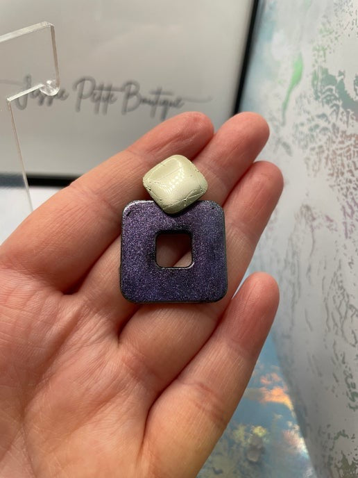 Hand holding a square-shaped earring with a gold top and purple base.