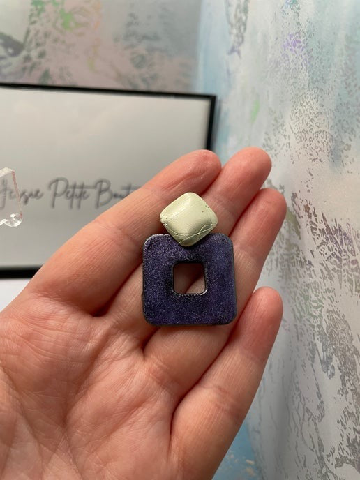 Hand holding a purple square-shaped earring against a textured wall.