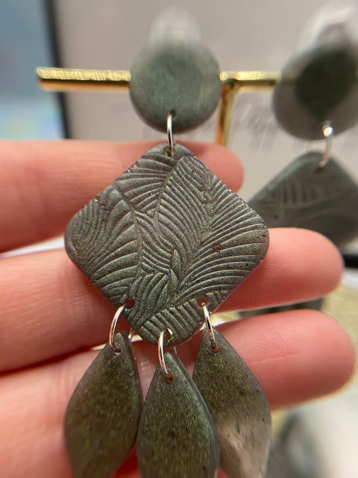 Earrings with textured green leaves held between fingers against a blurred background