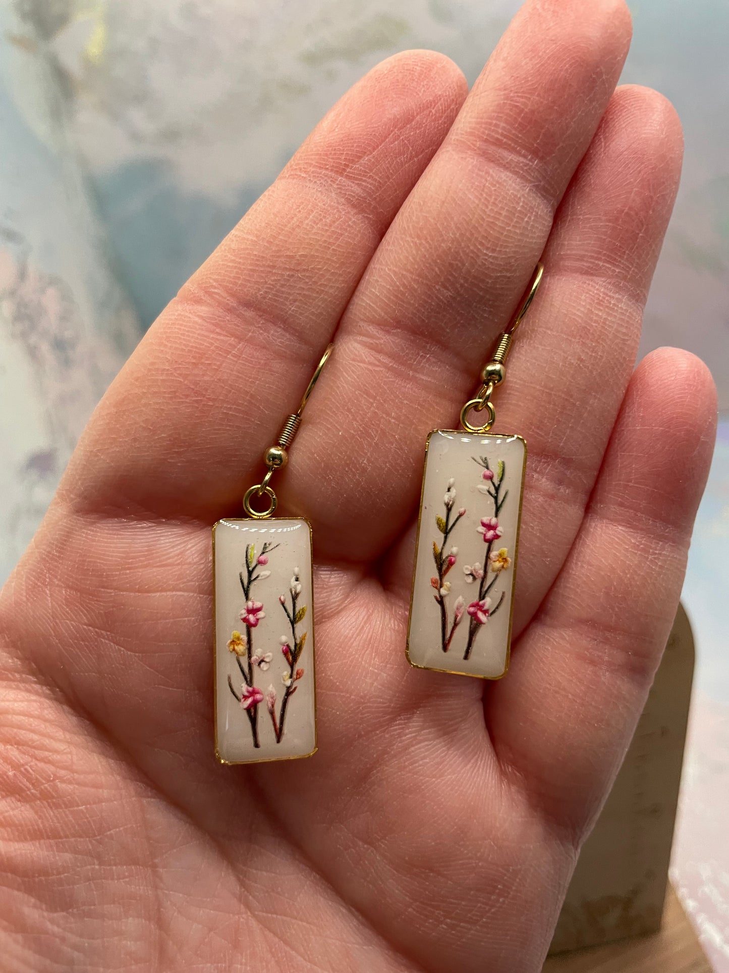 Decorative earrings with floral design held in a hand against a neutral background