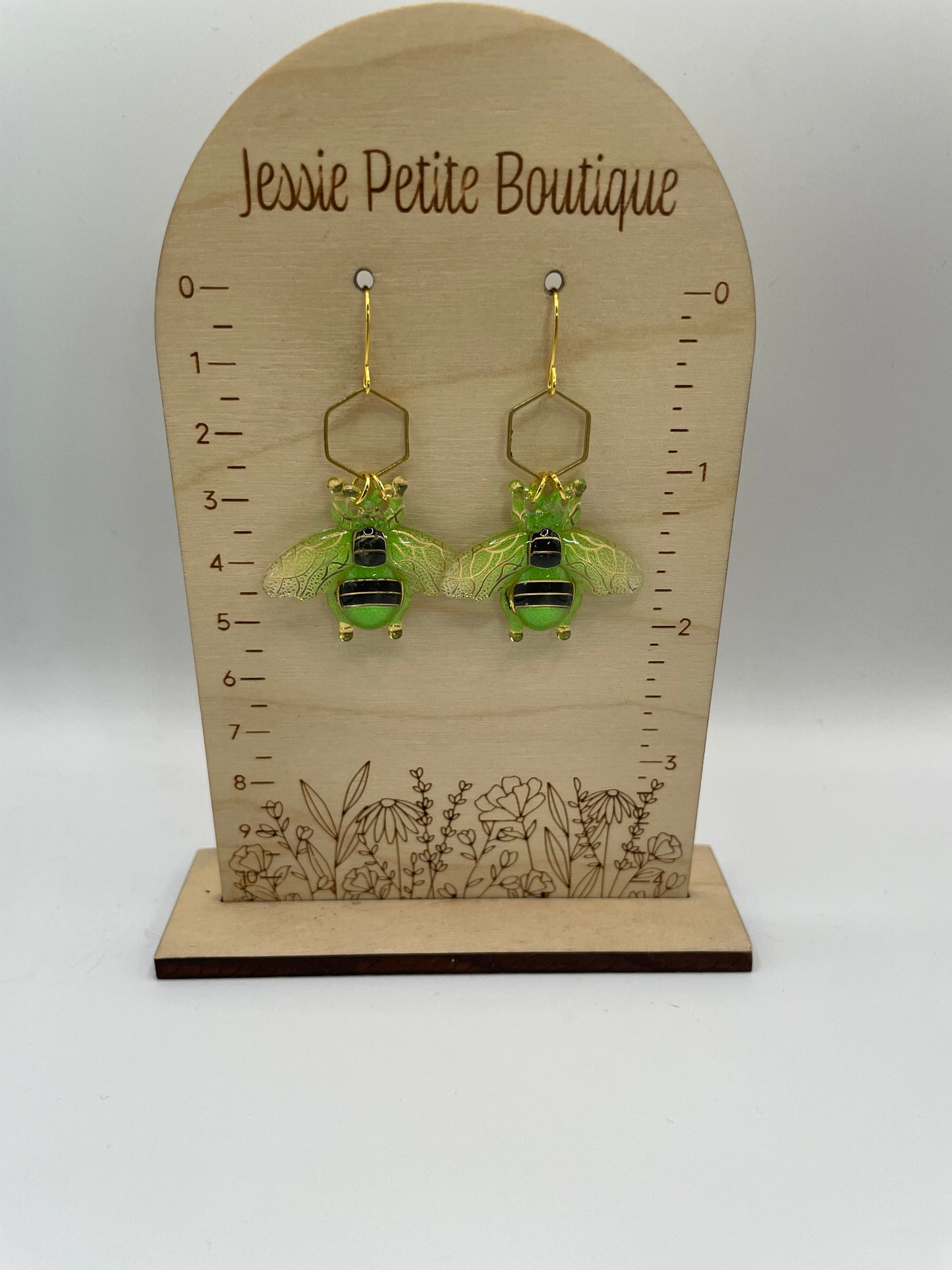Green bee-shaped earrings on a wooden display card with a ruler, branded 'Jessie Petite Boutique'.