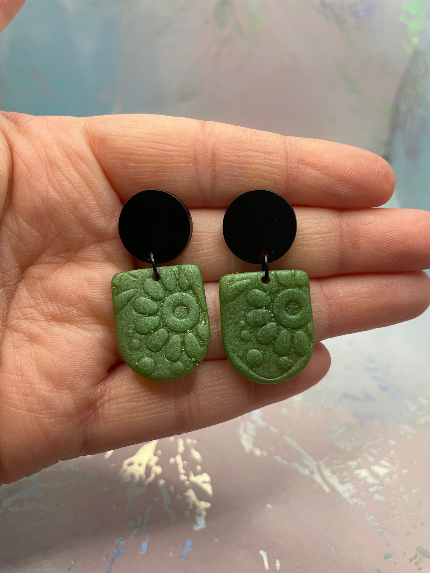 Green and black earrings held in a hand with a blurred background