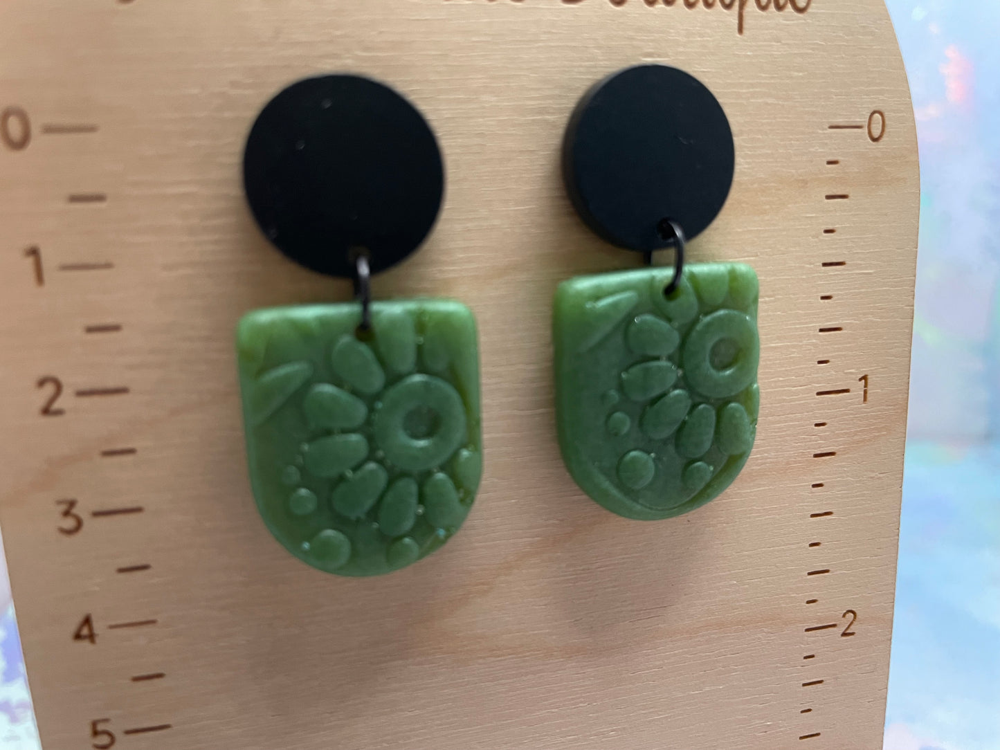 Green floral-patterned earrings with black circular tops on a wooden ruler.