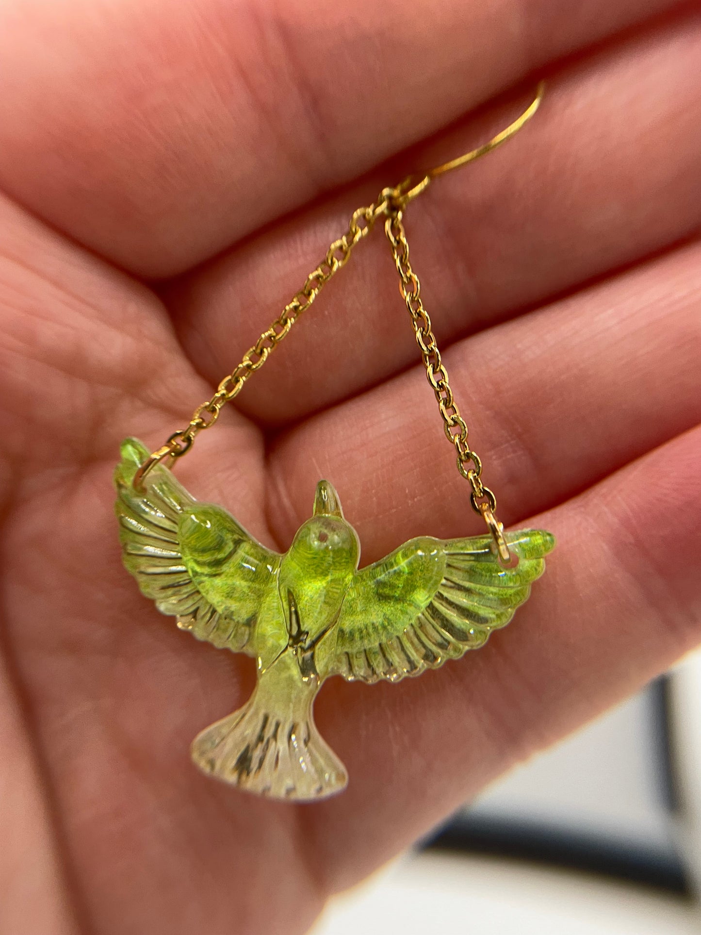 Green bird-shaped pendant on a gold chain held in a hand.