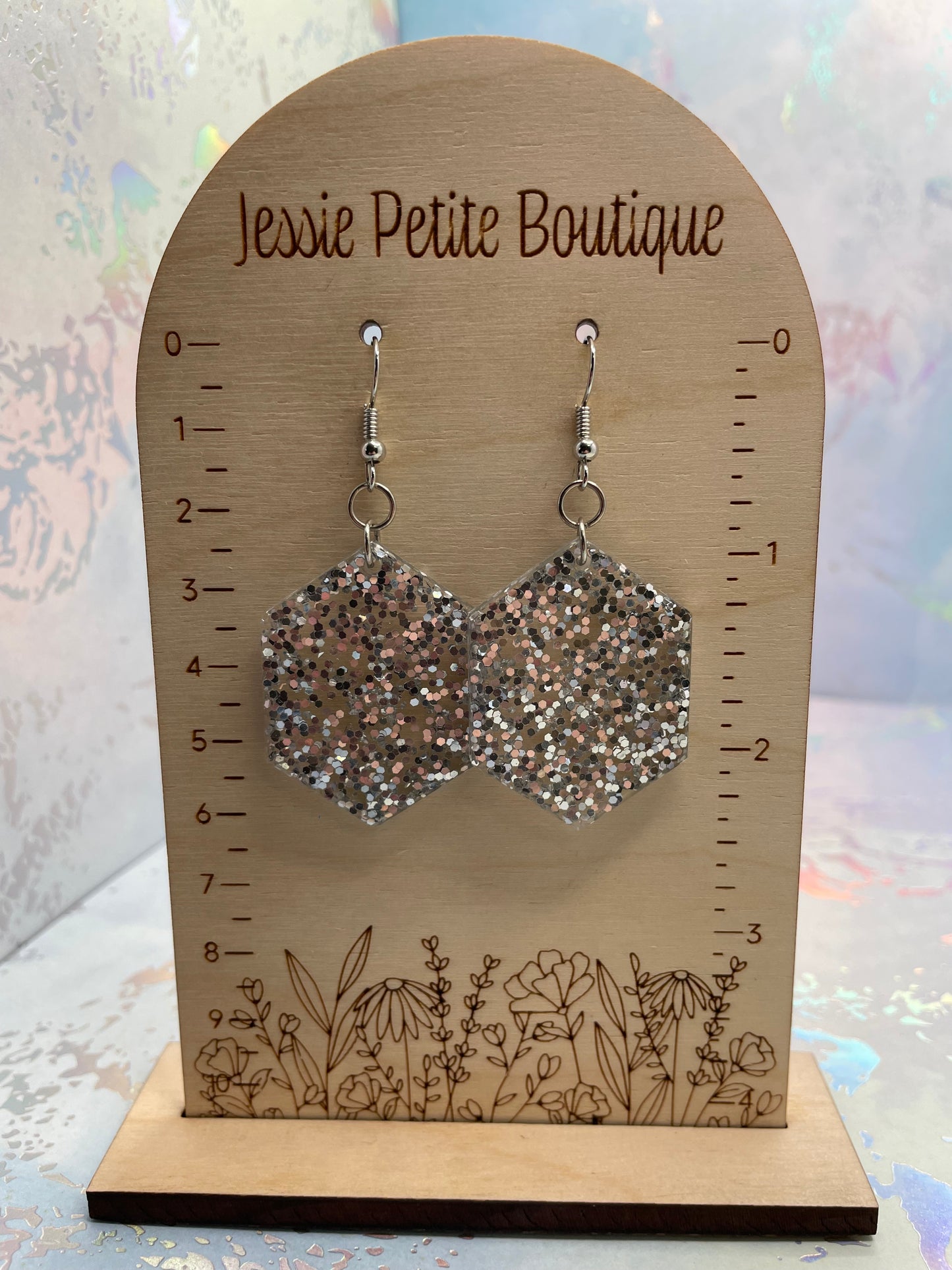 Pair of silver glittery earrings on a wooden measuring card with floral designs, branded 'Jessie Petite Boutique'.