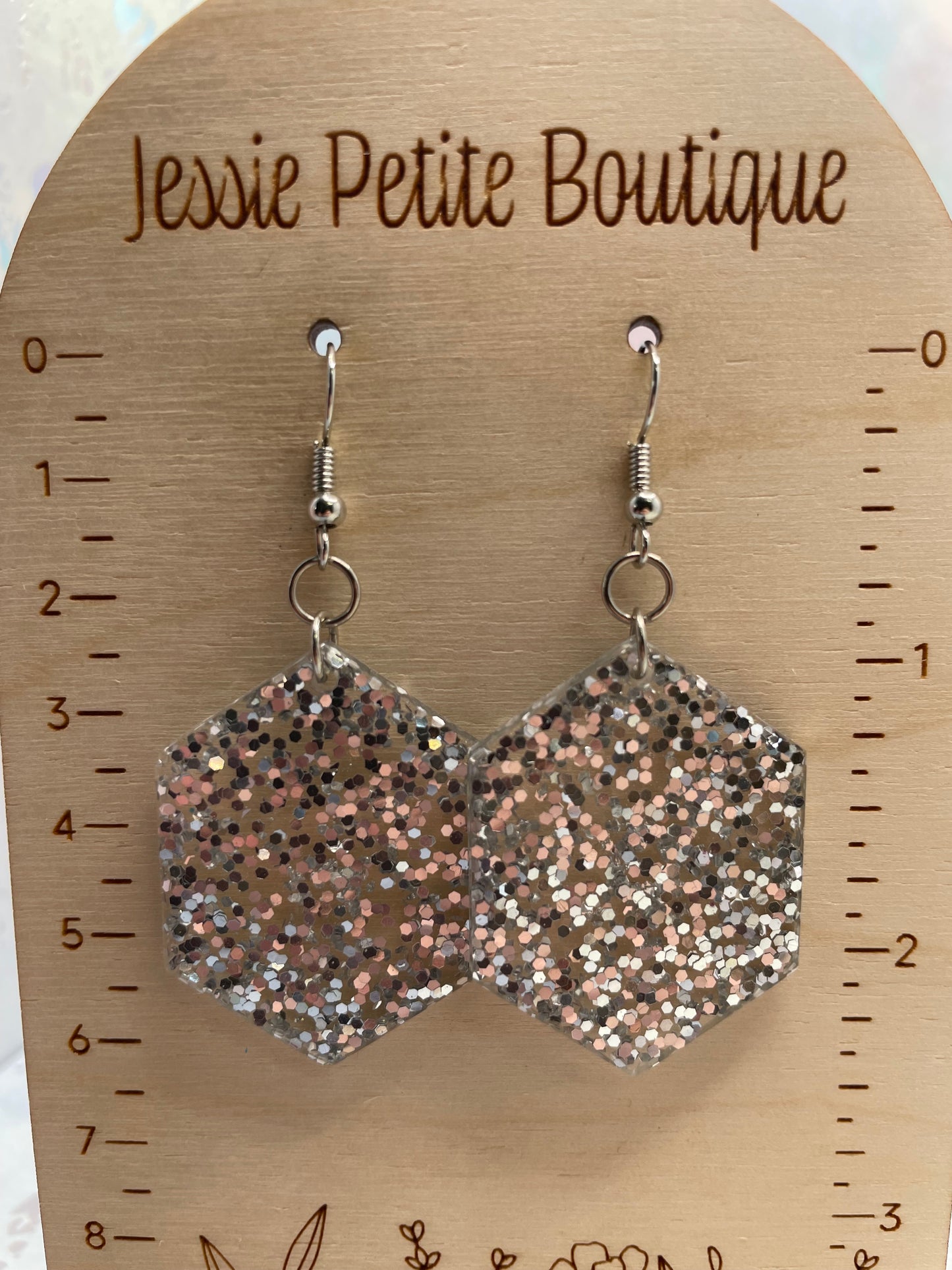 Hexagonal glittery earrings on a wooden measuring tool with 'Jessie Petite Boutique' branding.