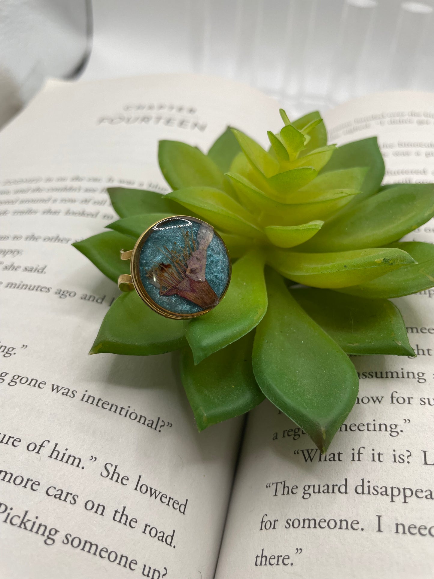 Succulent plant with a small round hydrangea flower on blue bezel gold adjustable ring on an open book.