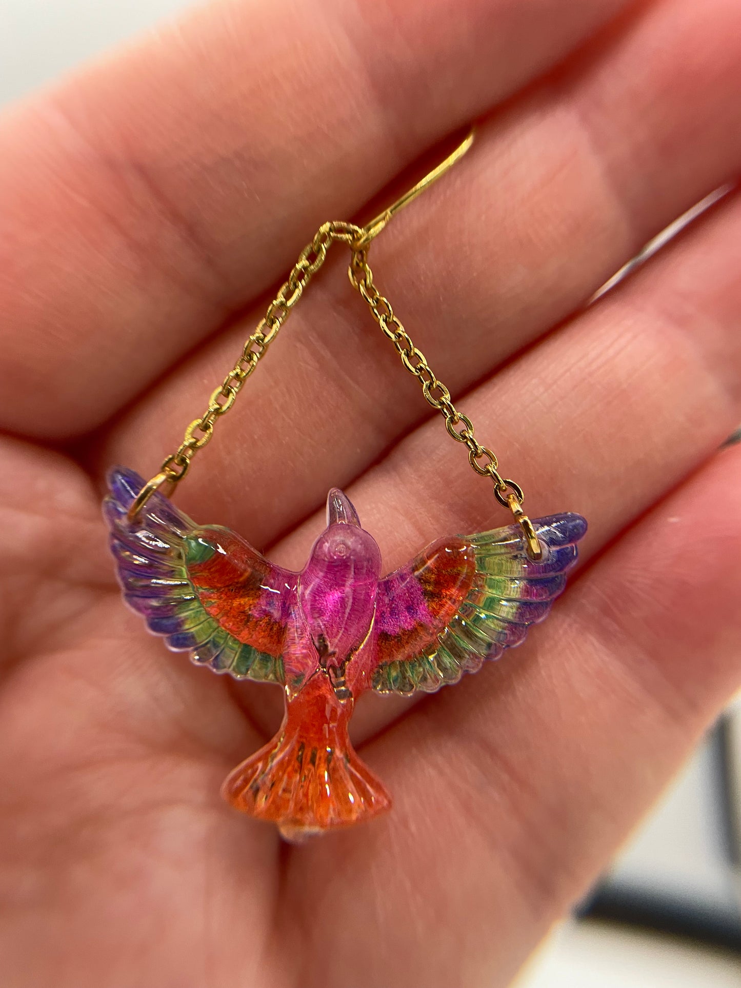 Colorful bird-shaped pendant on a gold chain held in a hand.
